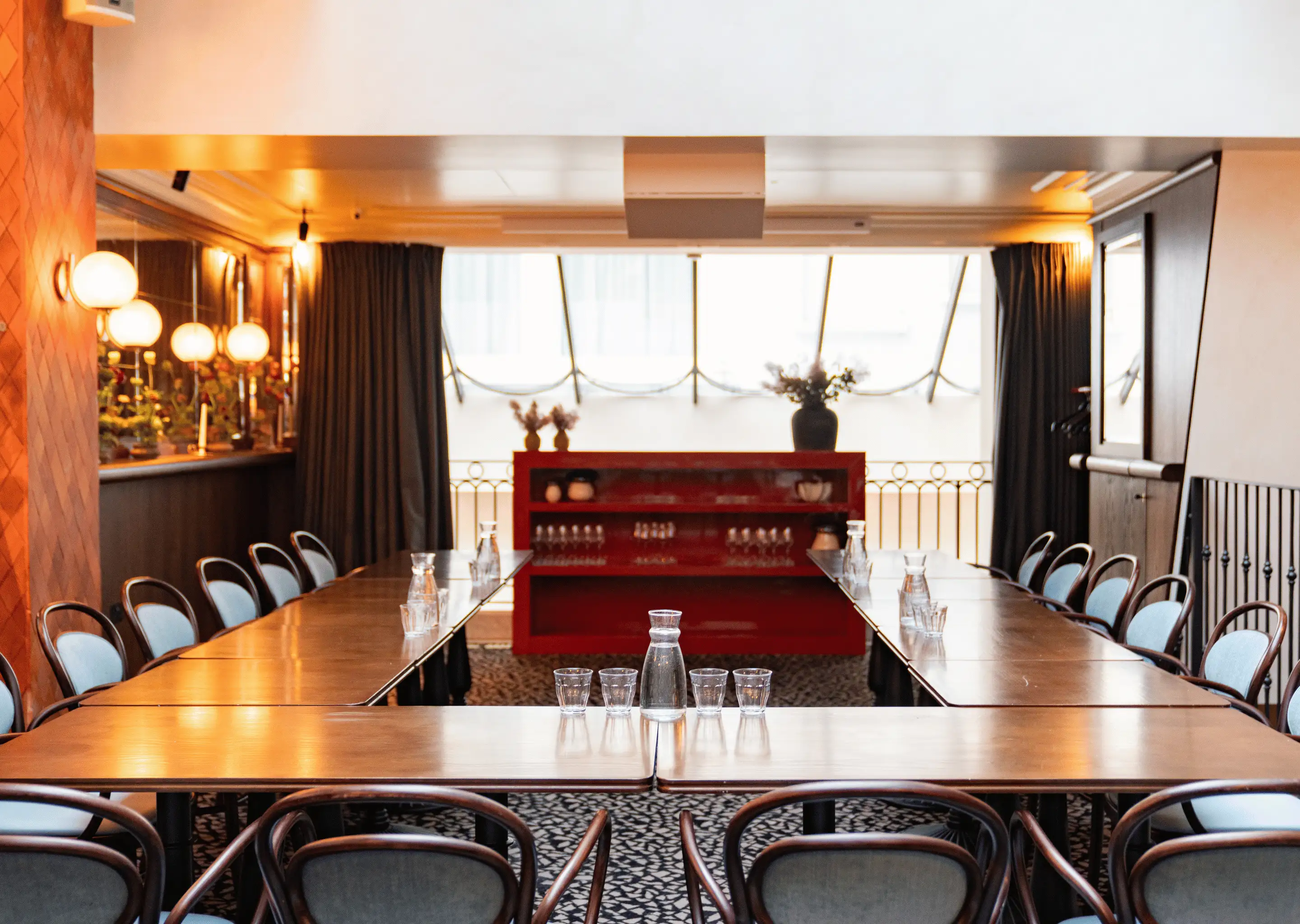Corporate meeting room with U-shaped wooden tables surrounded by chairs, glass pitchers, and glasses, featuring warm lighting and a red shelving unit with decorative items.
