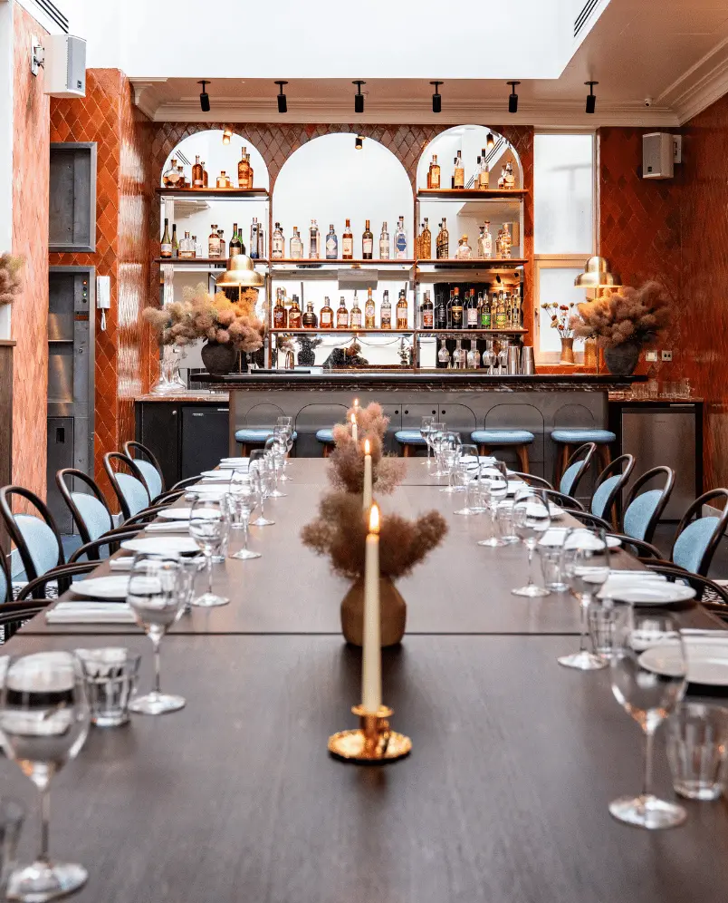 Elegant dining table set with glasses, plates, and candles facing a stylish bar with bottles and blue stools in a warm-toned room.