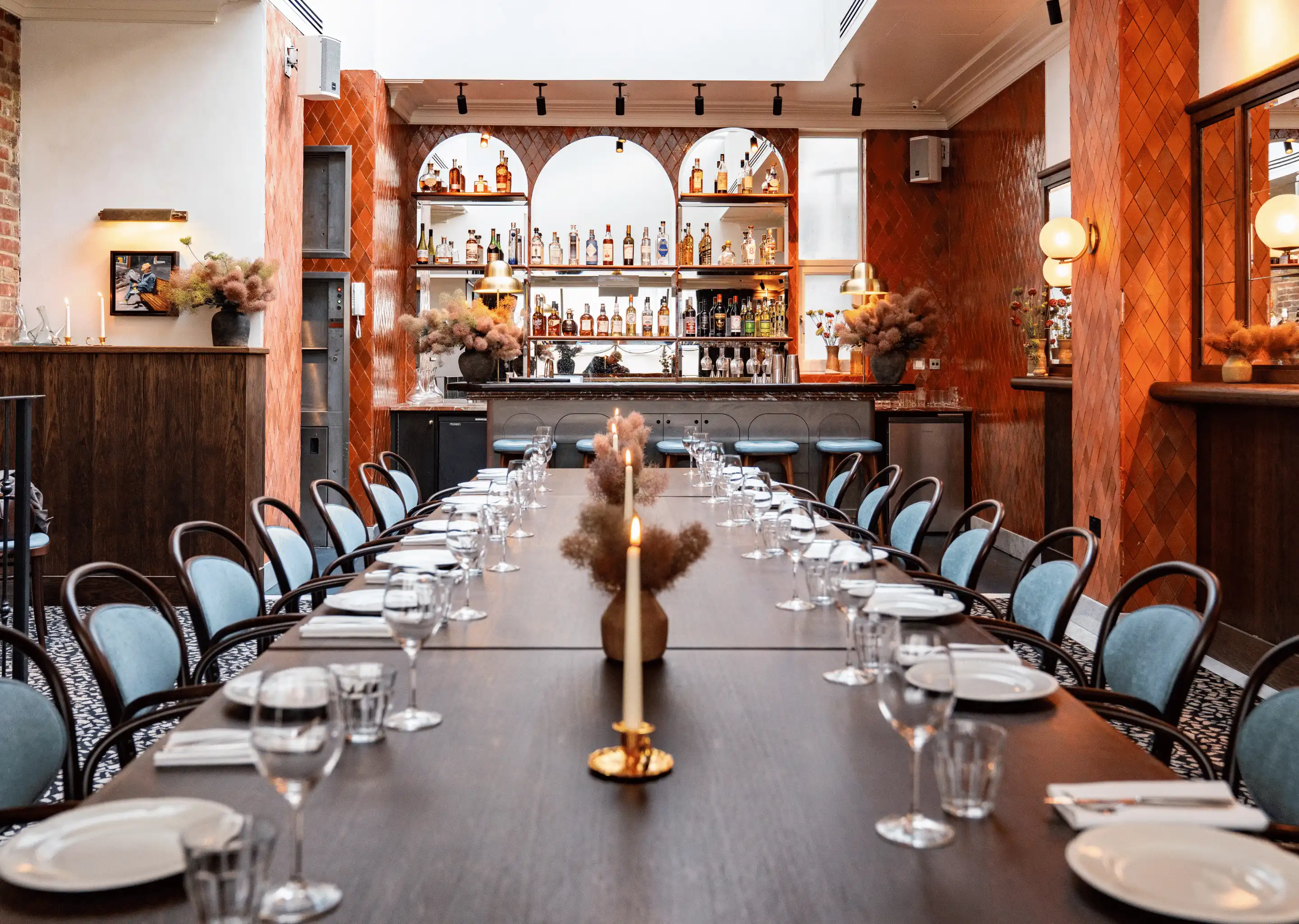 Long dining table set with plates, glasses, and candles in a stylish restaurant with a bar and orange tiled walls.