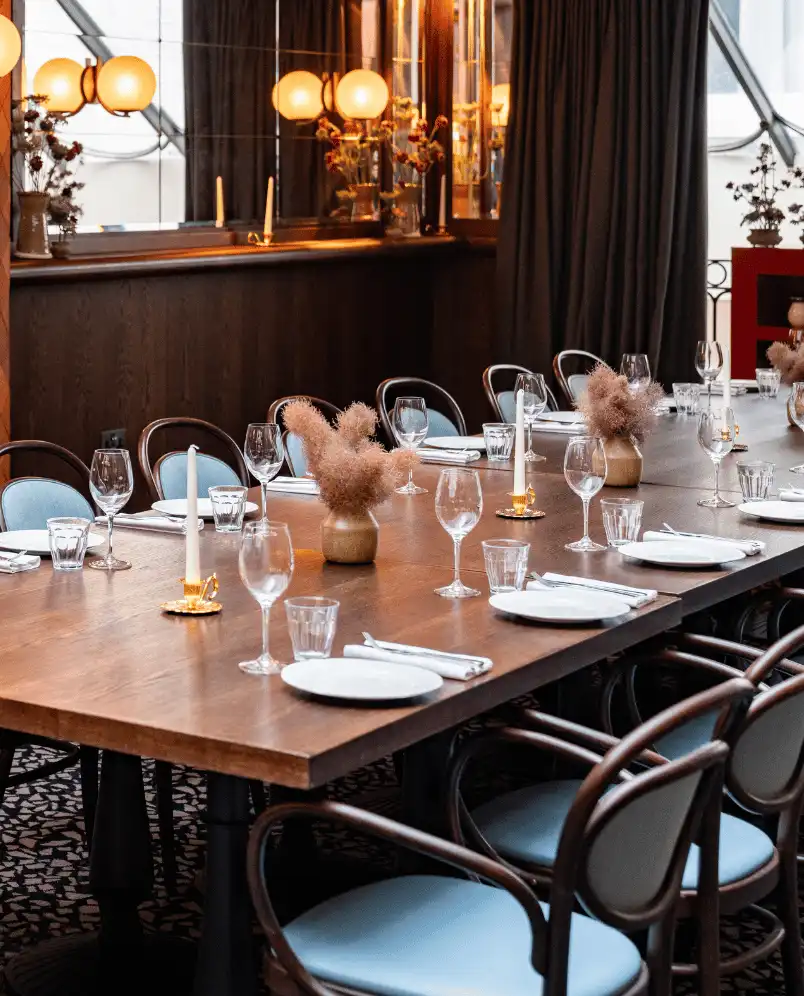 Long wooden dining table in a restaurant set with white plates, glassware, candles, and small vases with dried flowers.