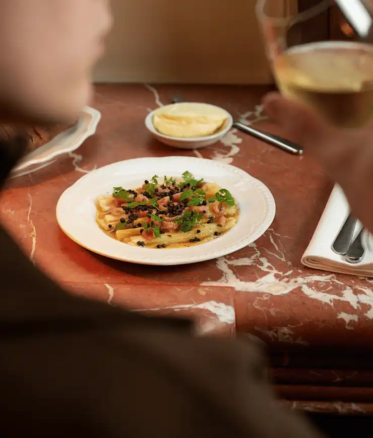Plate of pasta garnished with herbs and black pepper on a marble table next to a small dish of butter and a person holding a glass of white wine.