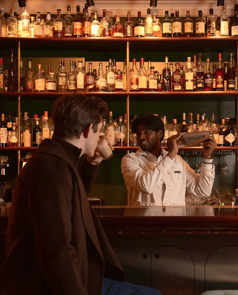 Bartender shaking a cocktail behind a bar with shelves filled with liquor bottles, while a seated man drinks.