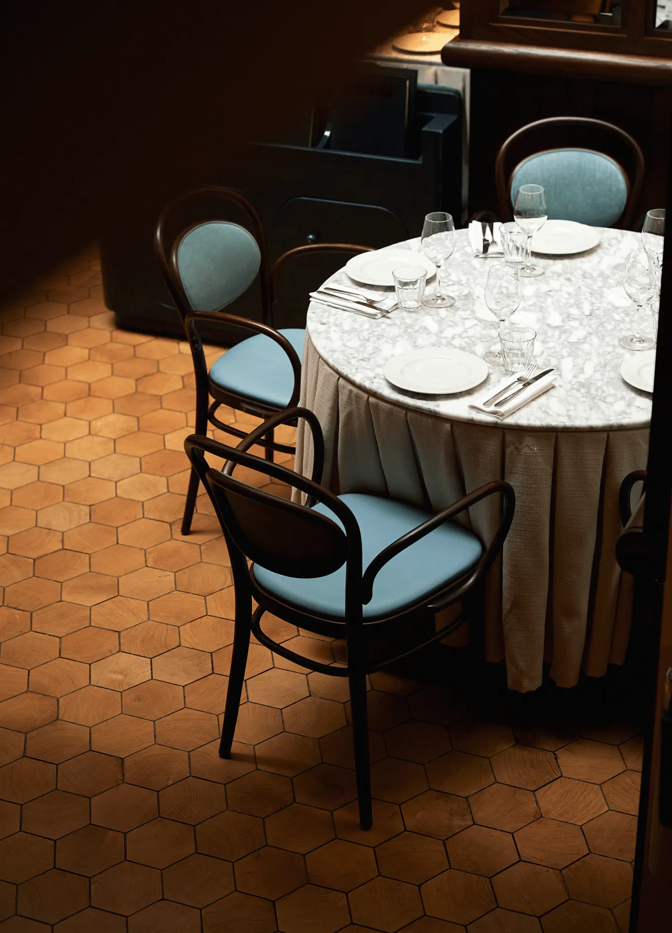 Round dining table with marble top set with plates, glasses, and silverware surrounded by wooden chairs with blue cushions on a hexagonal wood floor.