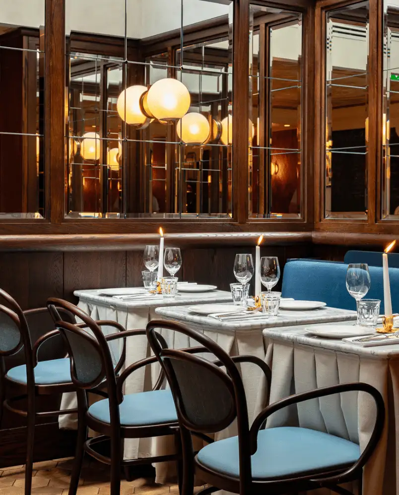 Elegant restaurant dining area with tables set with white candles, glassware, and plates, dark wood chairs with blue cushions, and mirrored walls reflecting round pendant lights.