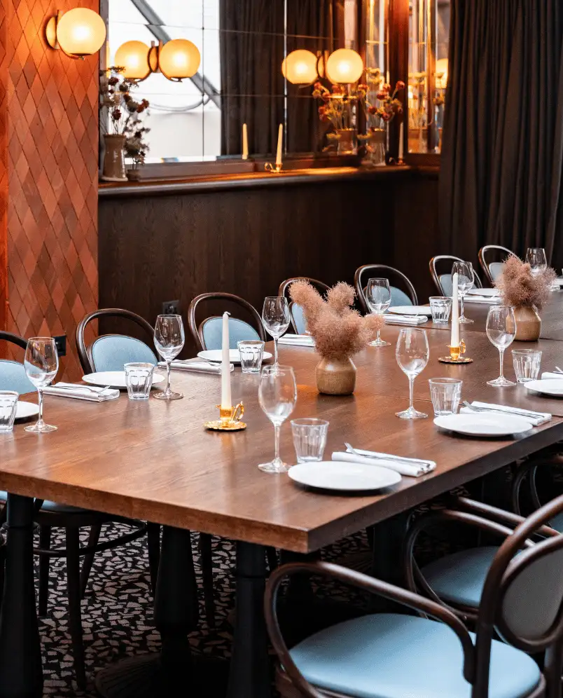 Long wooden dining table set with plates, wine glasses, water tumblers, candles, and vases with dried flowers in a warm-lit restaurant.