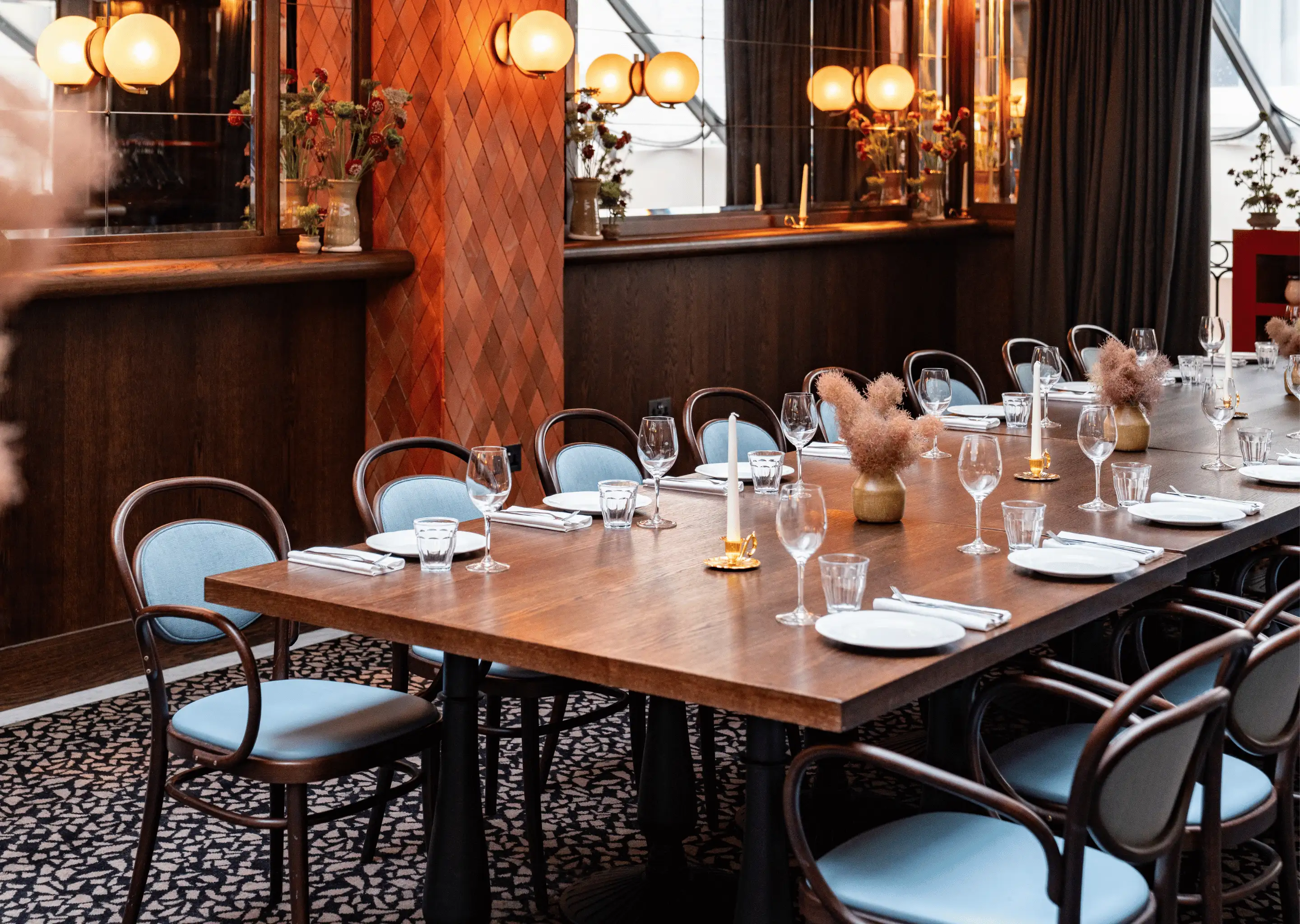 Long wooden dining table set with plates, glasses, white napkins, candles, and small floral centerpieces in a warmly lit restaurant.