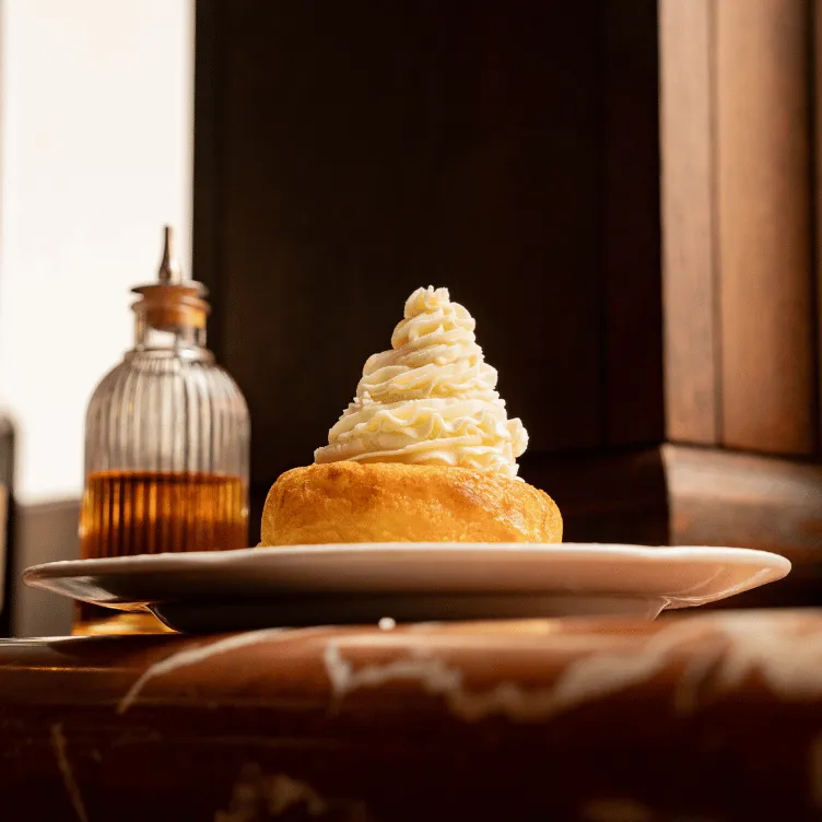 Close-up of a dessert with a swirl of whipped cream on a round pastry on a white plate and a glass bottle with amber liquid in the background.