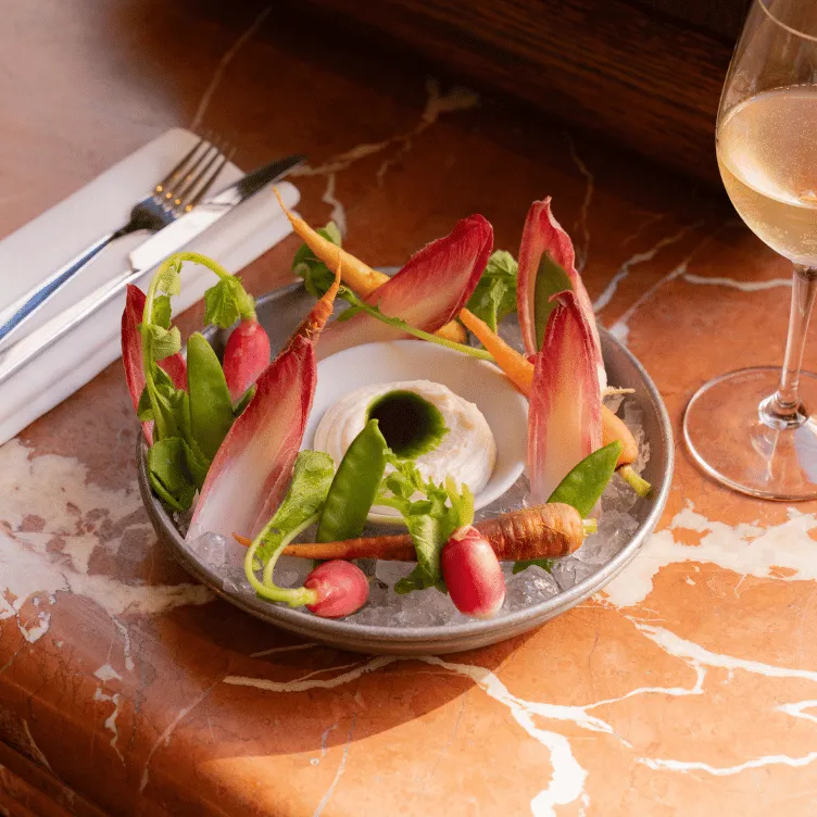 Plate of fresh vegetables and dip served on ice, with a glass of white wine and cutlery on a folded napkin on a marble table.