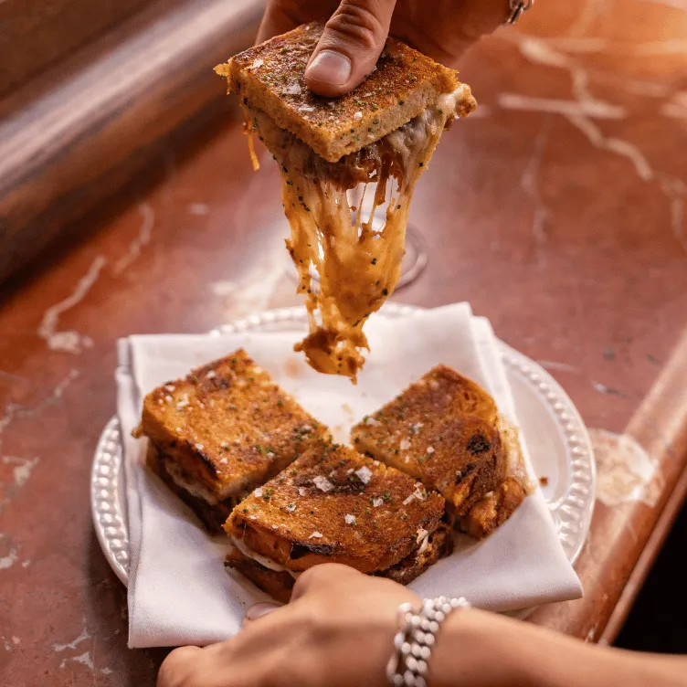 Close-up of a hand lifting a toasted sandwich quarter with melted cheese stretching, with three more sandwich quarters on a white napkin on a plate.