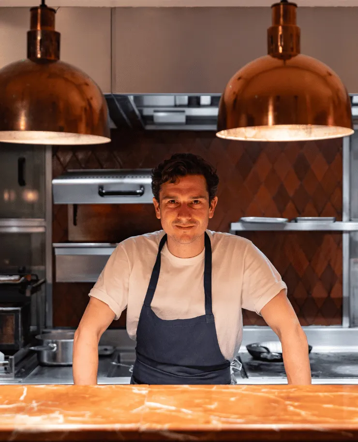 Chef wearing a white shirt and dark apron standing behind a counter in a warmly lit kitchen.