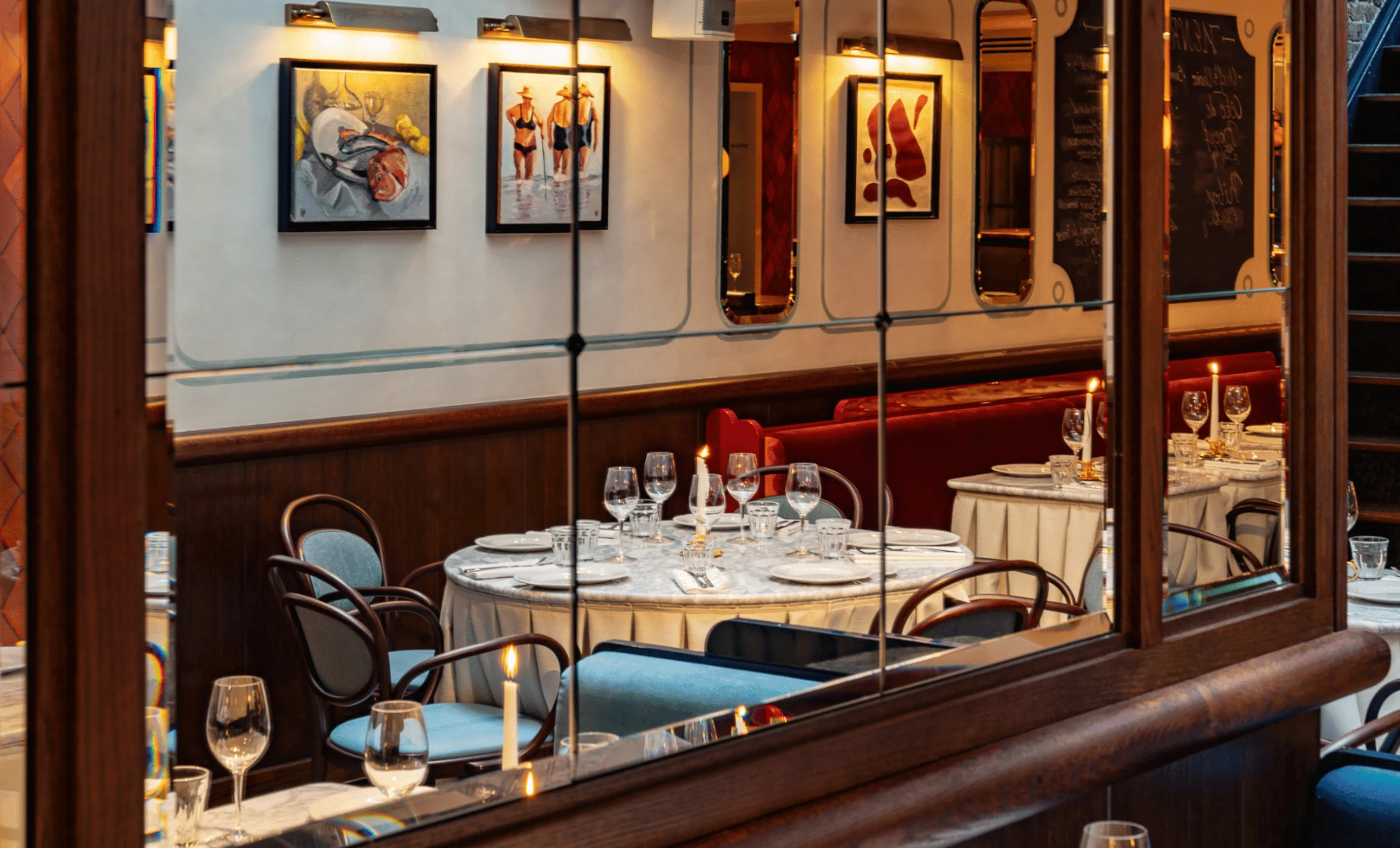 Interior of a restaurant with a round table set with plates, glasses, and lit candles, surrounded by blue and brown chairs, with framed artwork on the wall and a red cushioned bench in the background.