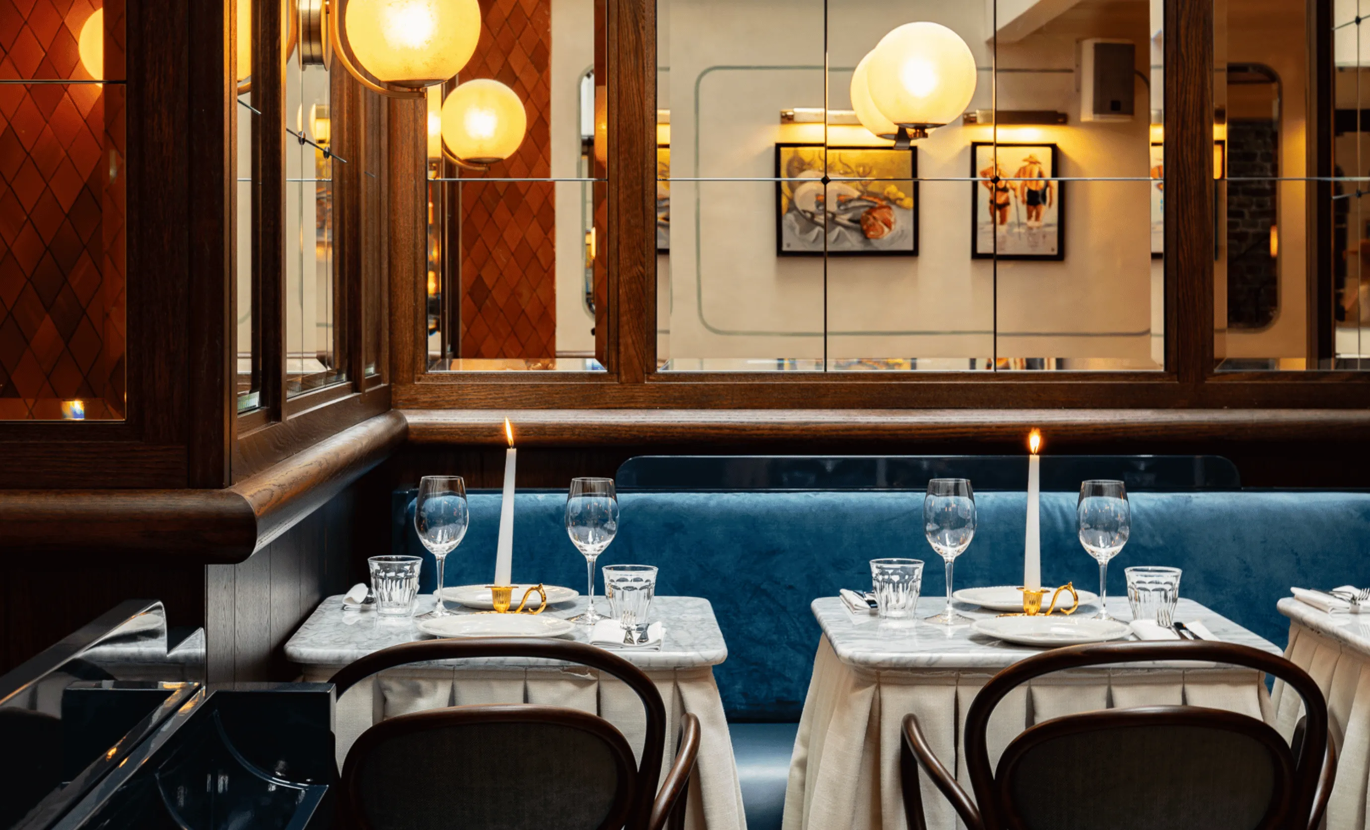 Elegant restaurant seating with marble tables, blue velvet bench, lit candles, and mirrored walls reflecting warm lighting and framed art.