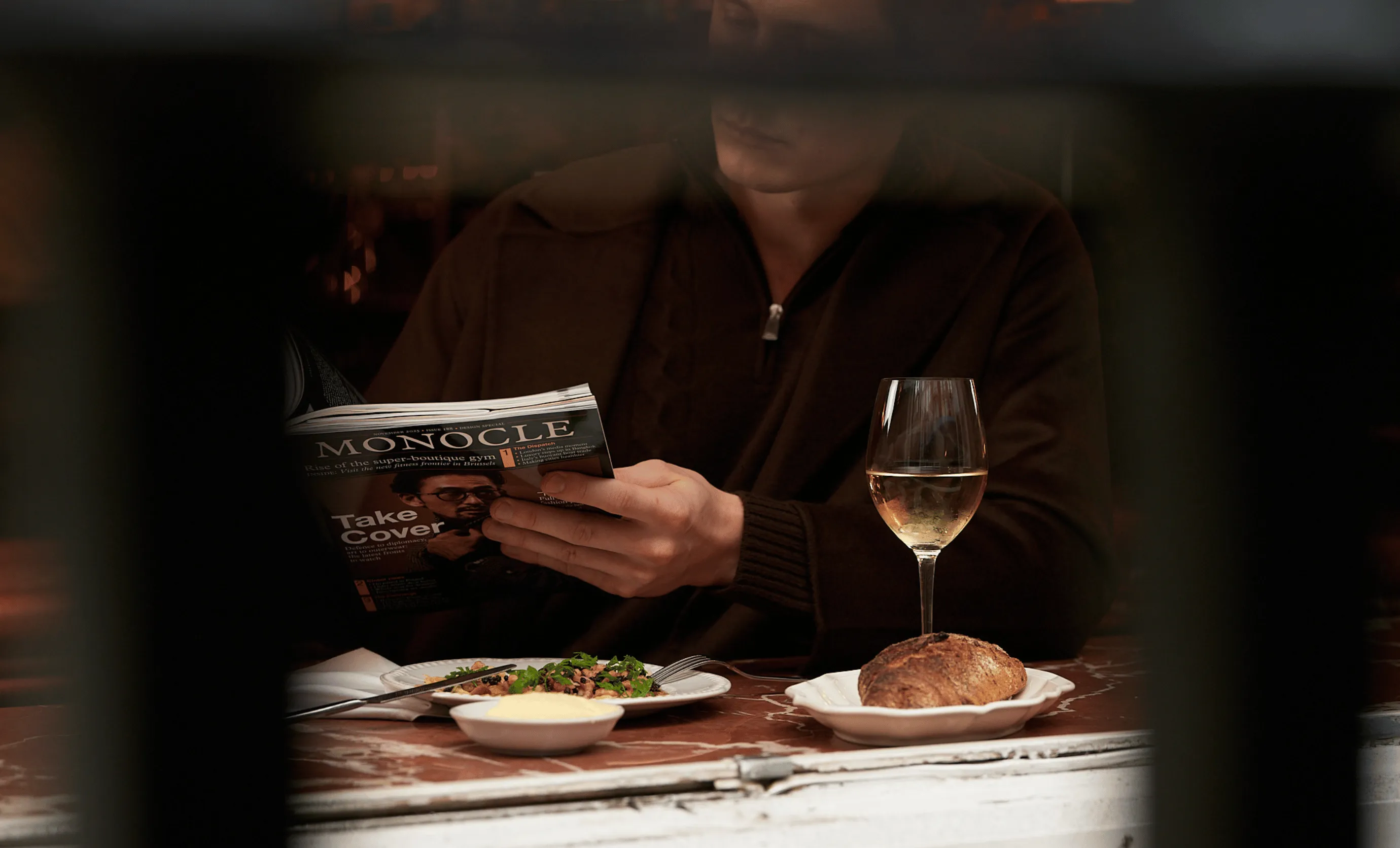 Person reading Monocle magazine while sitting at a table with a plate of food, a bread roll, a small dish, and a glass of white wine.