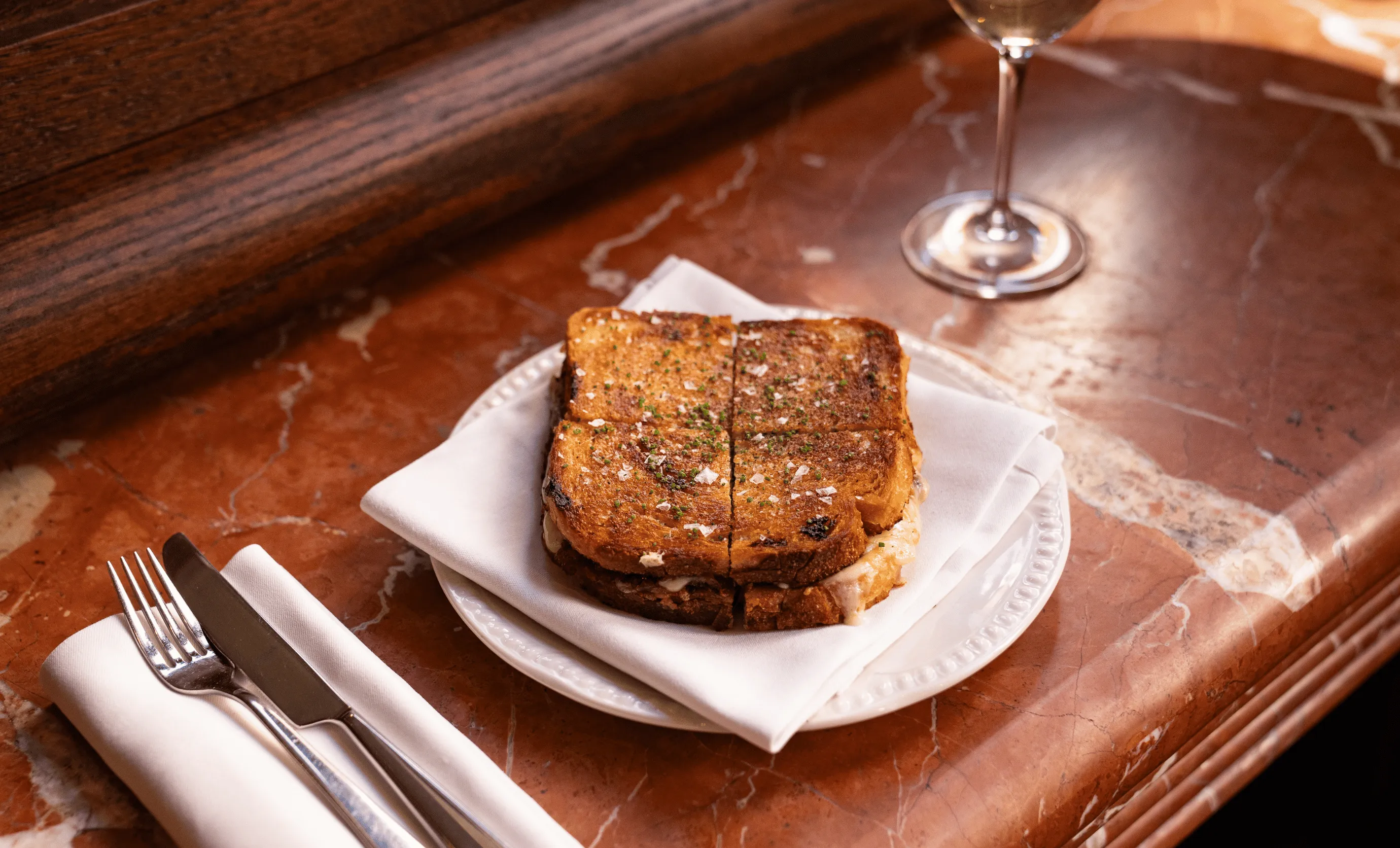 Grilled cheese sandwich cut in half, served on a white plate with a napkin on a marble table with a fork, knife, and glass of water nearby.