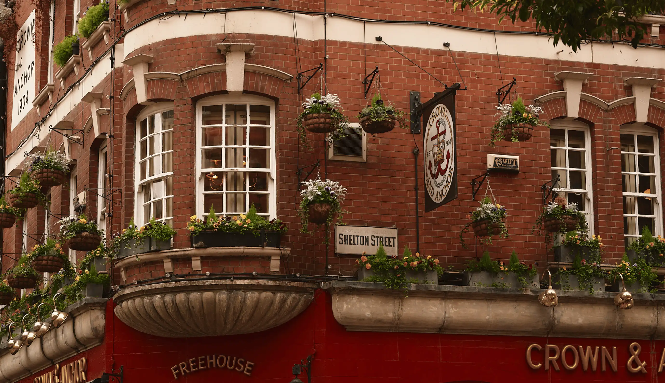 Red brick building corner with hanging flower baskets, windows, and a Crown and Anchor pub sign on Shelton Street.