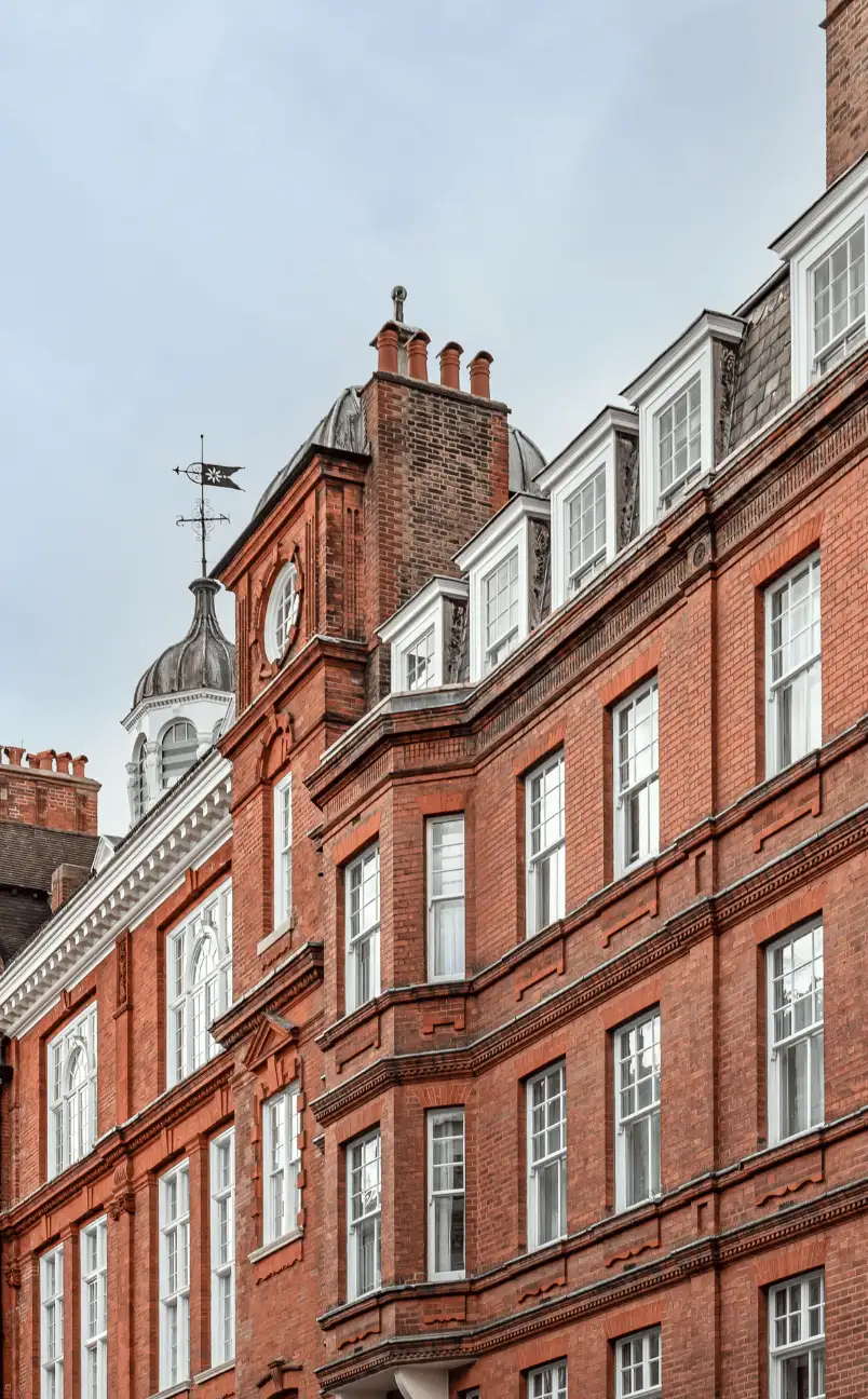 Red brick building with multiple white-framed windows and a cupola featuring a weather vane under a cloudy sky.