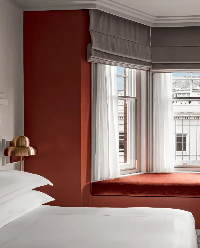 Hotel room corner with white bed linens, a brass bedside lamp, burnt orange wall, and a window seat with cushioned bench under gray roman shades.