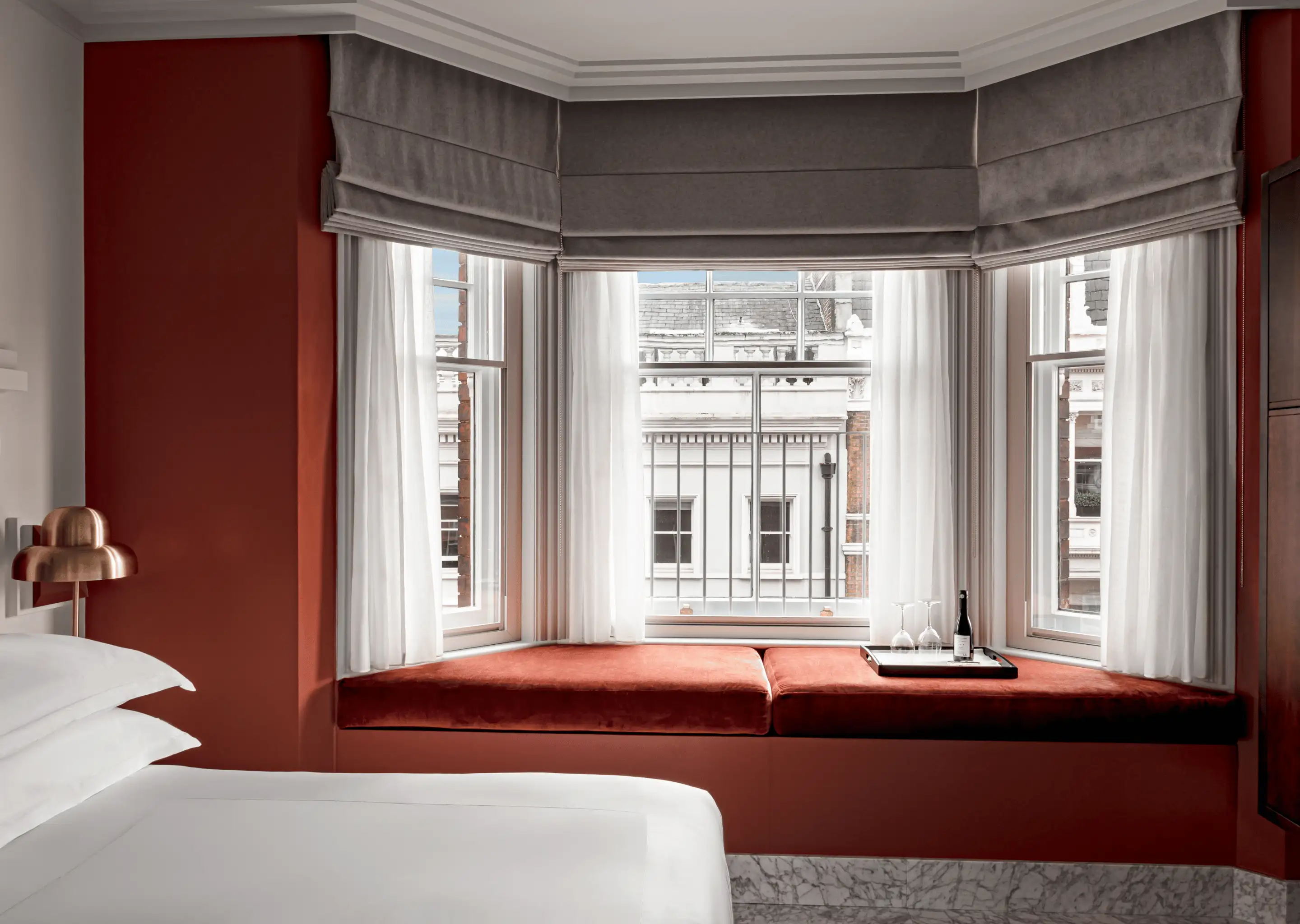 Cozy hotel room corner with a white bed, red cushioned window seat, sheer curtains, and a tray with wine and glasses.