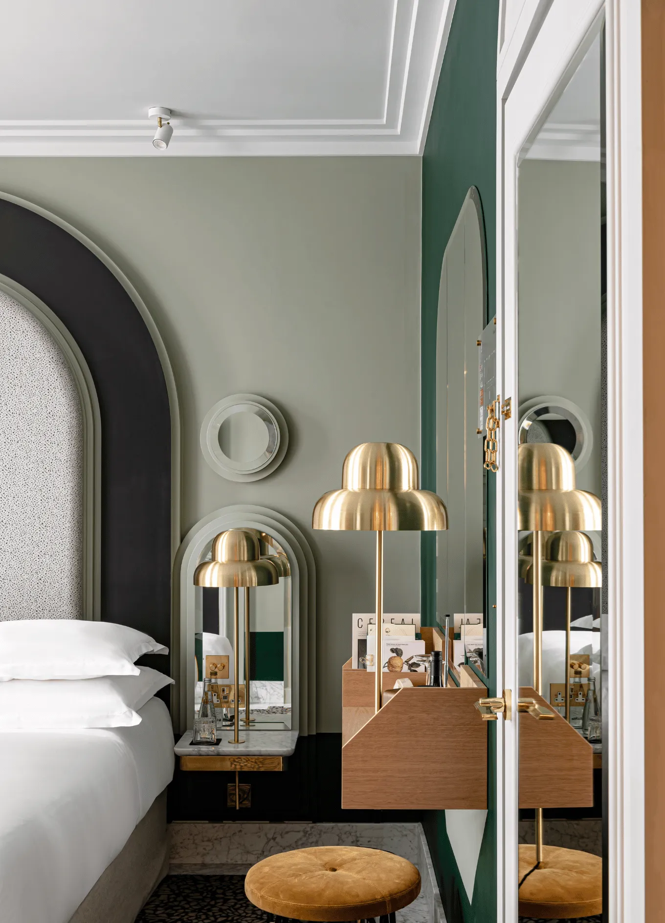 Modern hotel bedroom corner with white bed, layered arched mirrors, brass lamp, wooden floating desk, and round mustard stool.