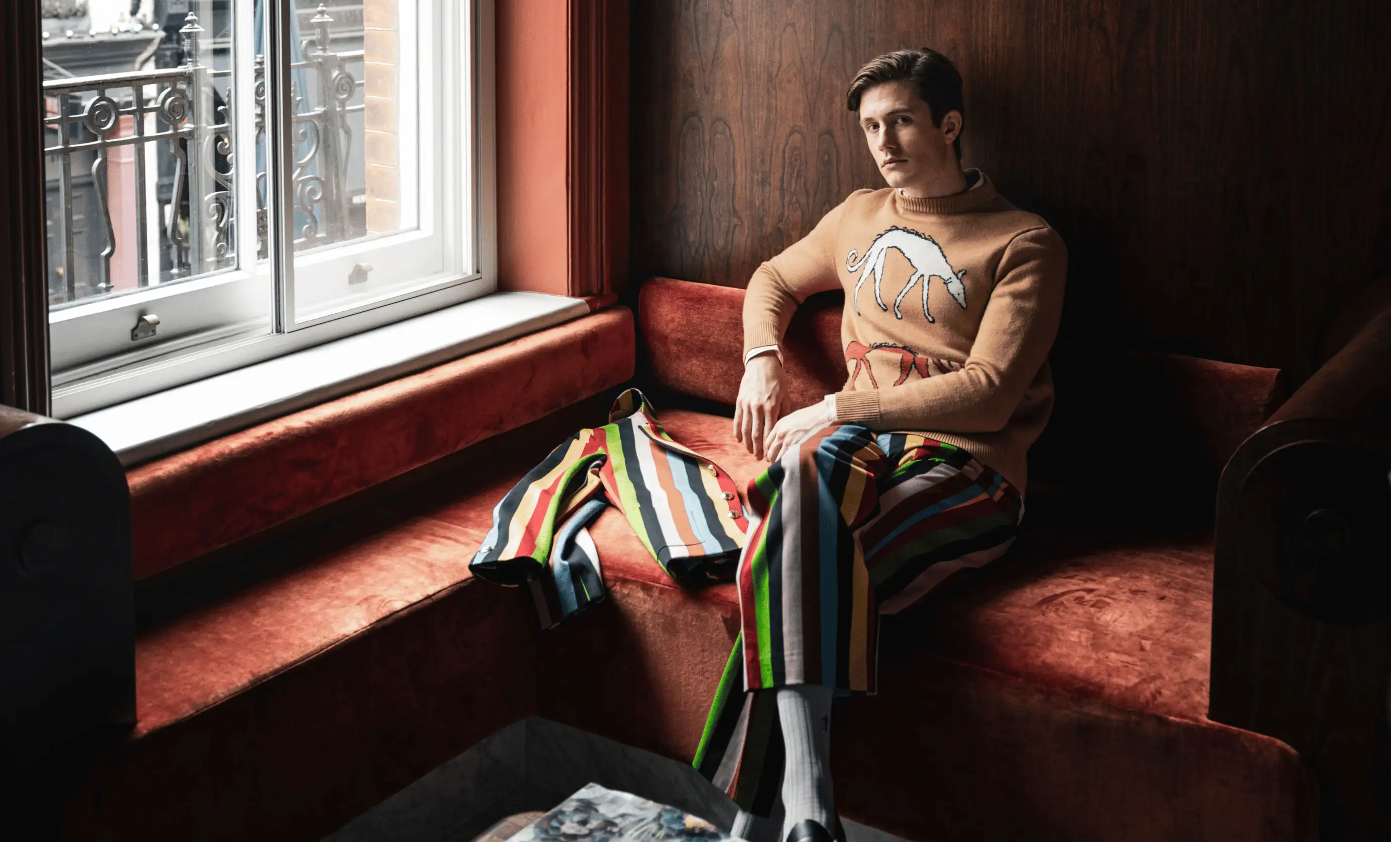Man wearing a tan sweater with a white dog graphic and multicolored striped pants sitting on a red velvet corner bench next to a window.