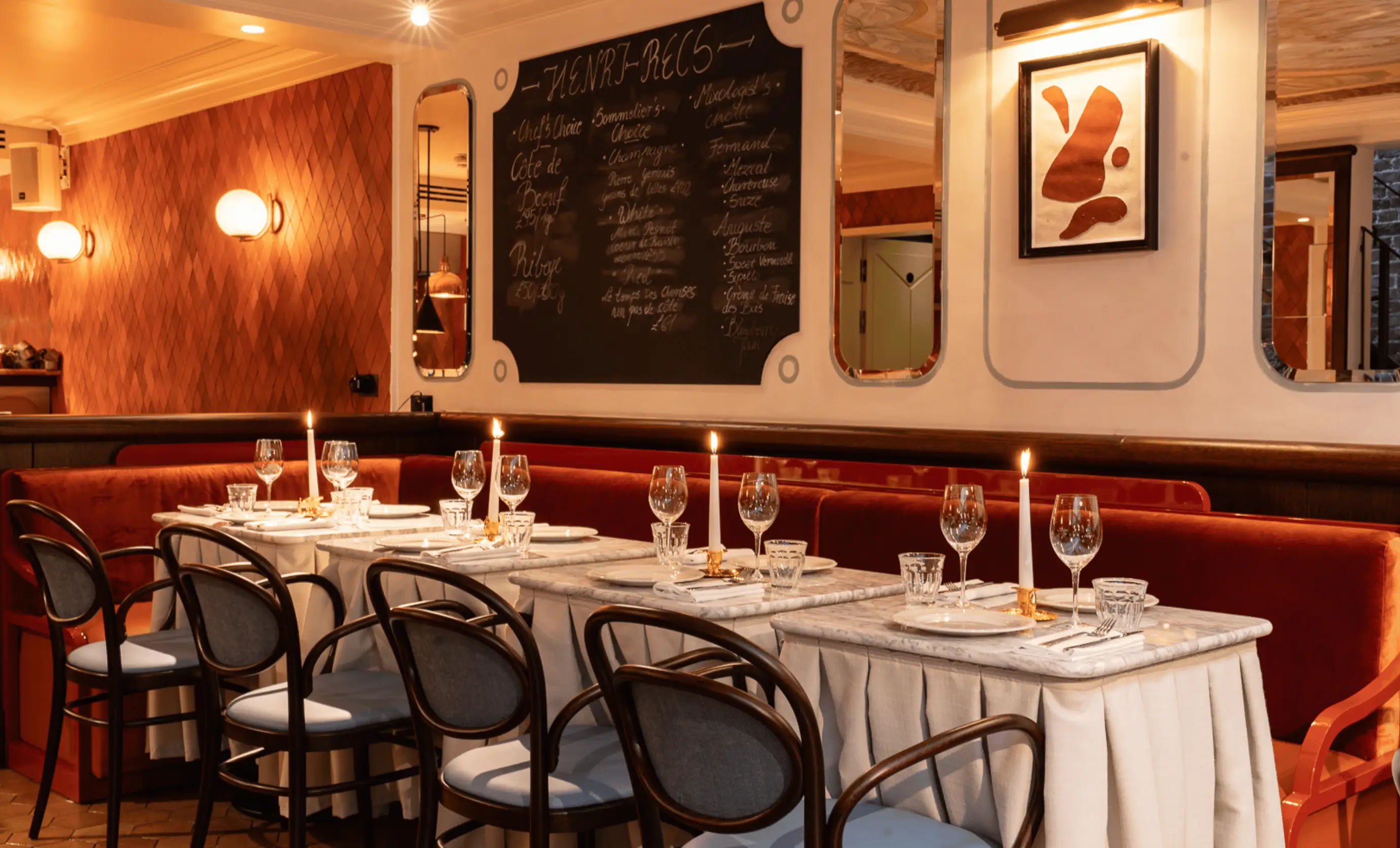 Cozy restaurant interior with marble tables, red velvet bench seating, lit candles, and chairs with blue cushions under warm lighting.