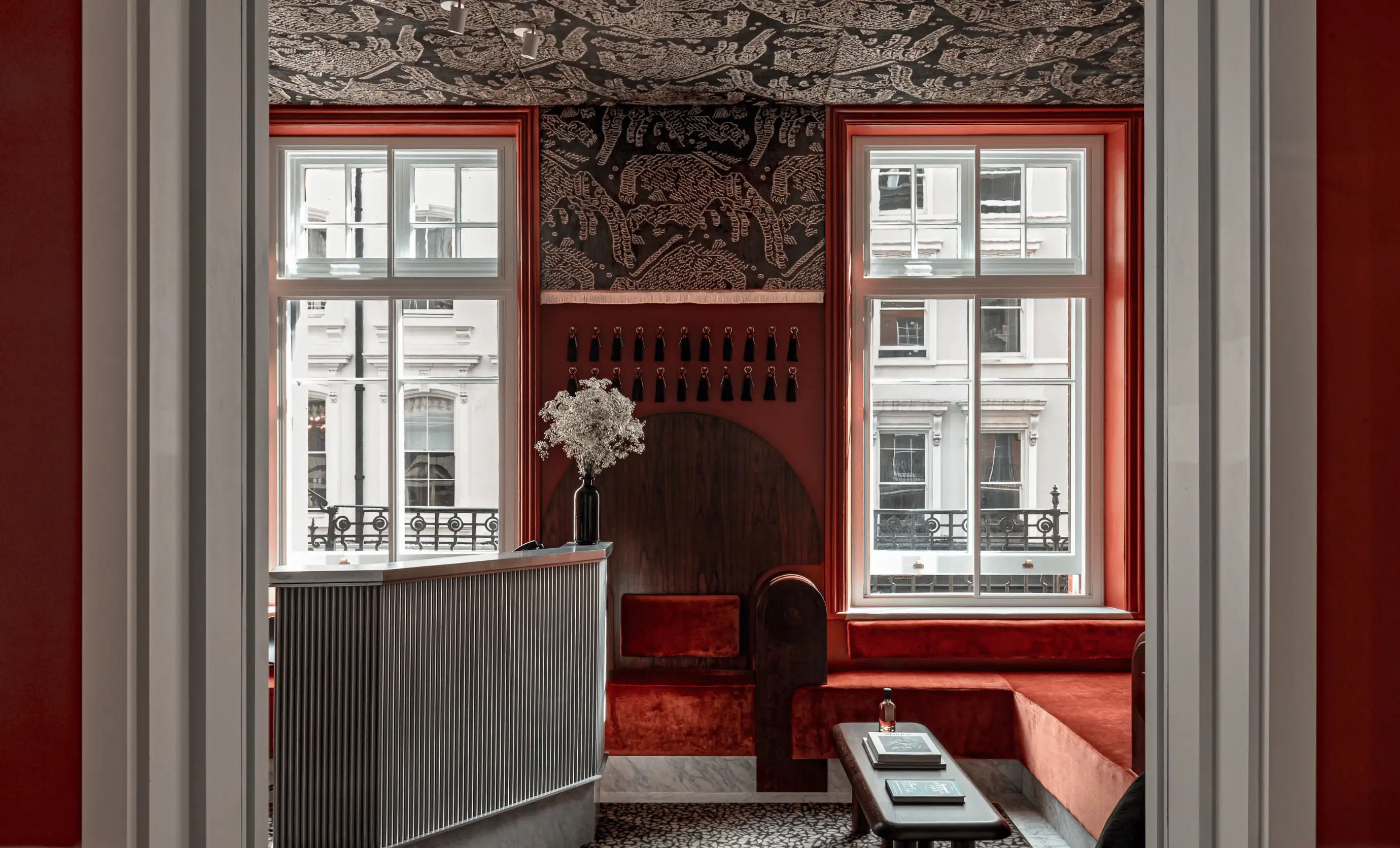 Hotel reception area with red velvet seating, two large windows, black and white patterned ceiling, and a check-in desk with a vase of white flowers.