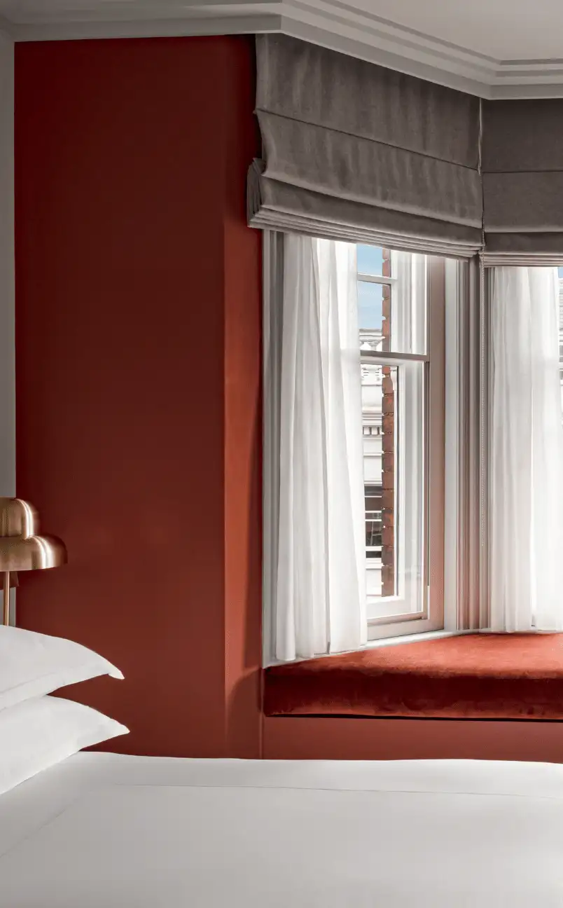 Cozy corner in hotel room with rust-colored walls, window seat with cushion, sheer white curtains, and part of a white bed with pillows.