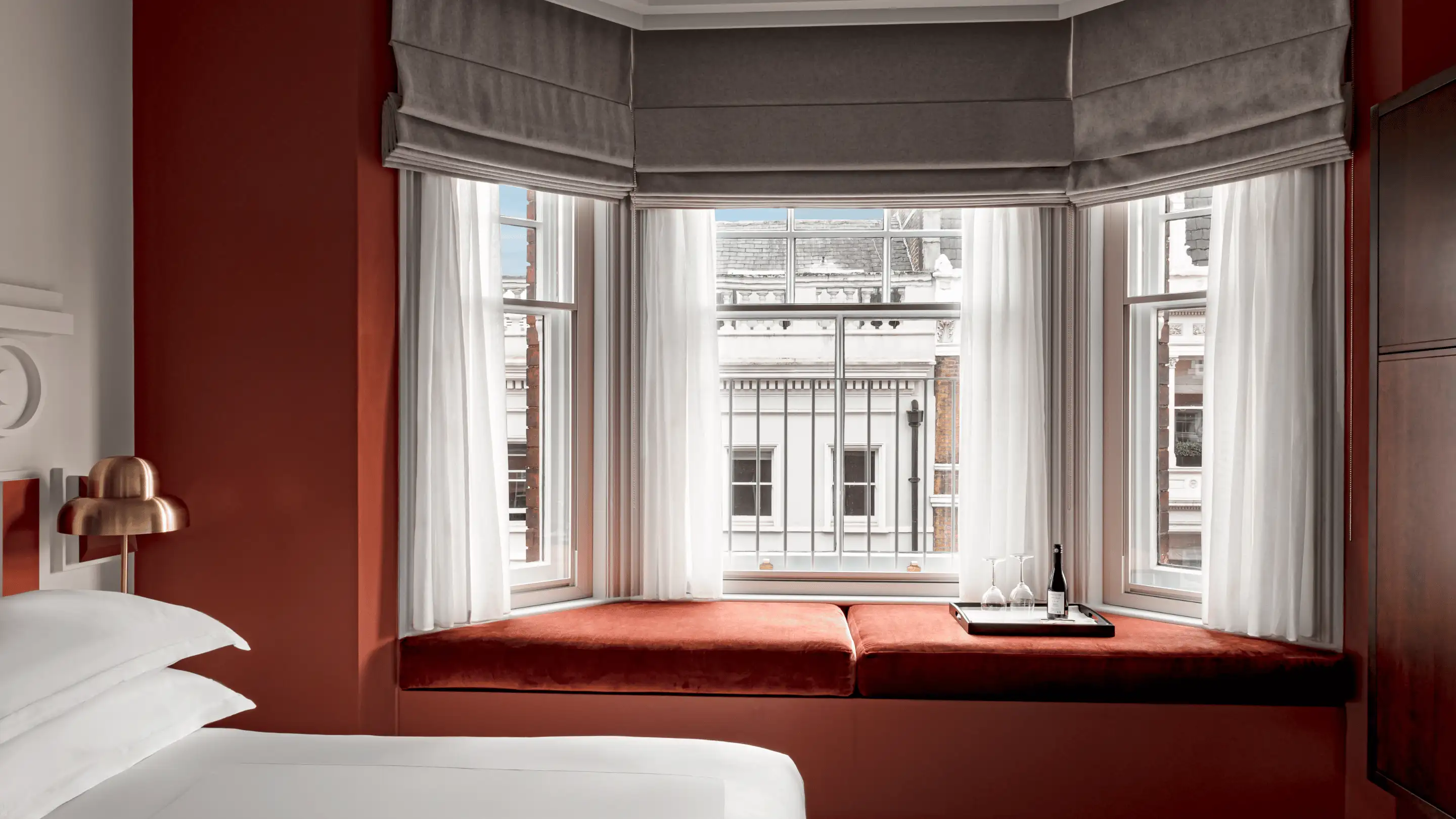 Cozy hotel room featuring a white bed, copper lamp, and cushioned window seat with a tray holding wine glasses and a bottle.