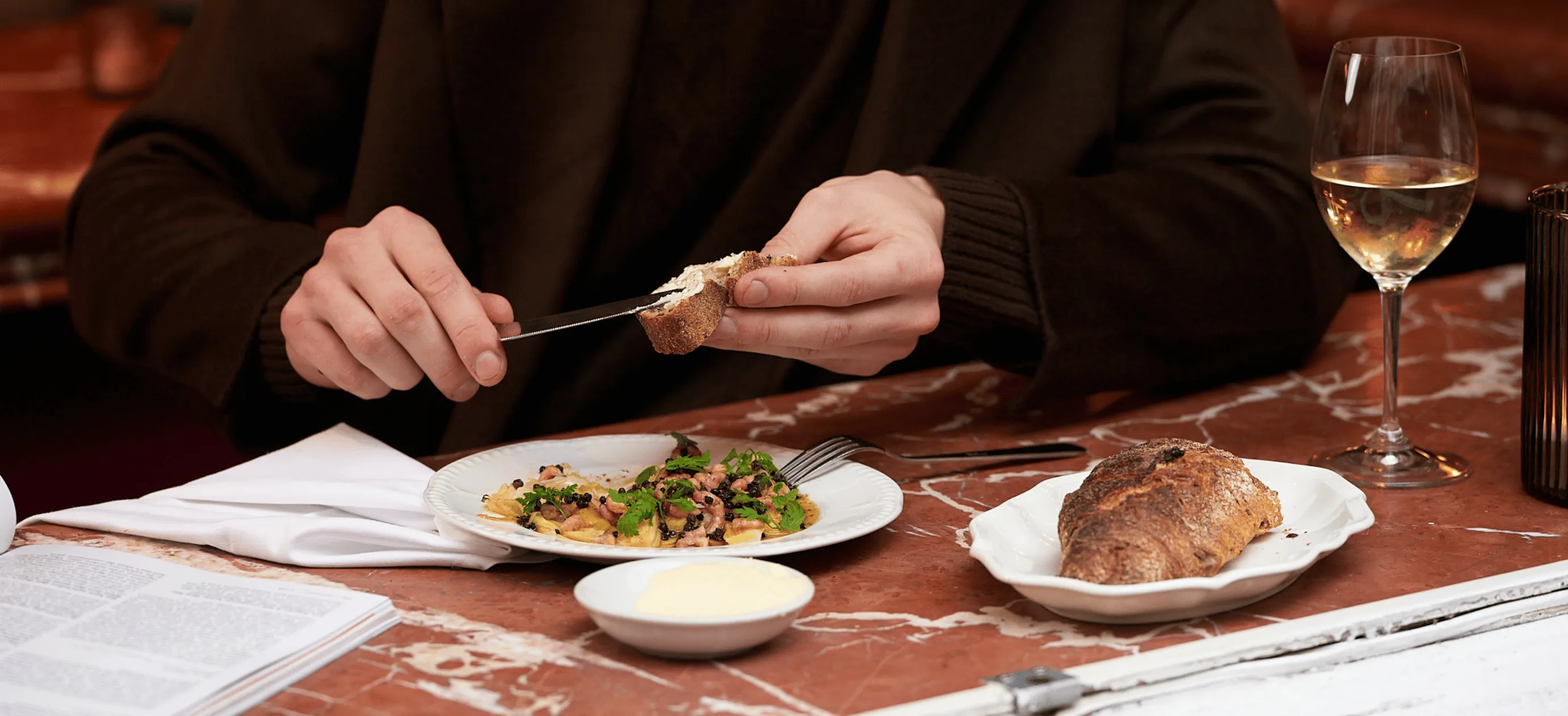 Person spreading butter on a piece of bread at a table with a plate of food, a bread roll, a glass of white wine, and an open book.