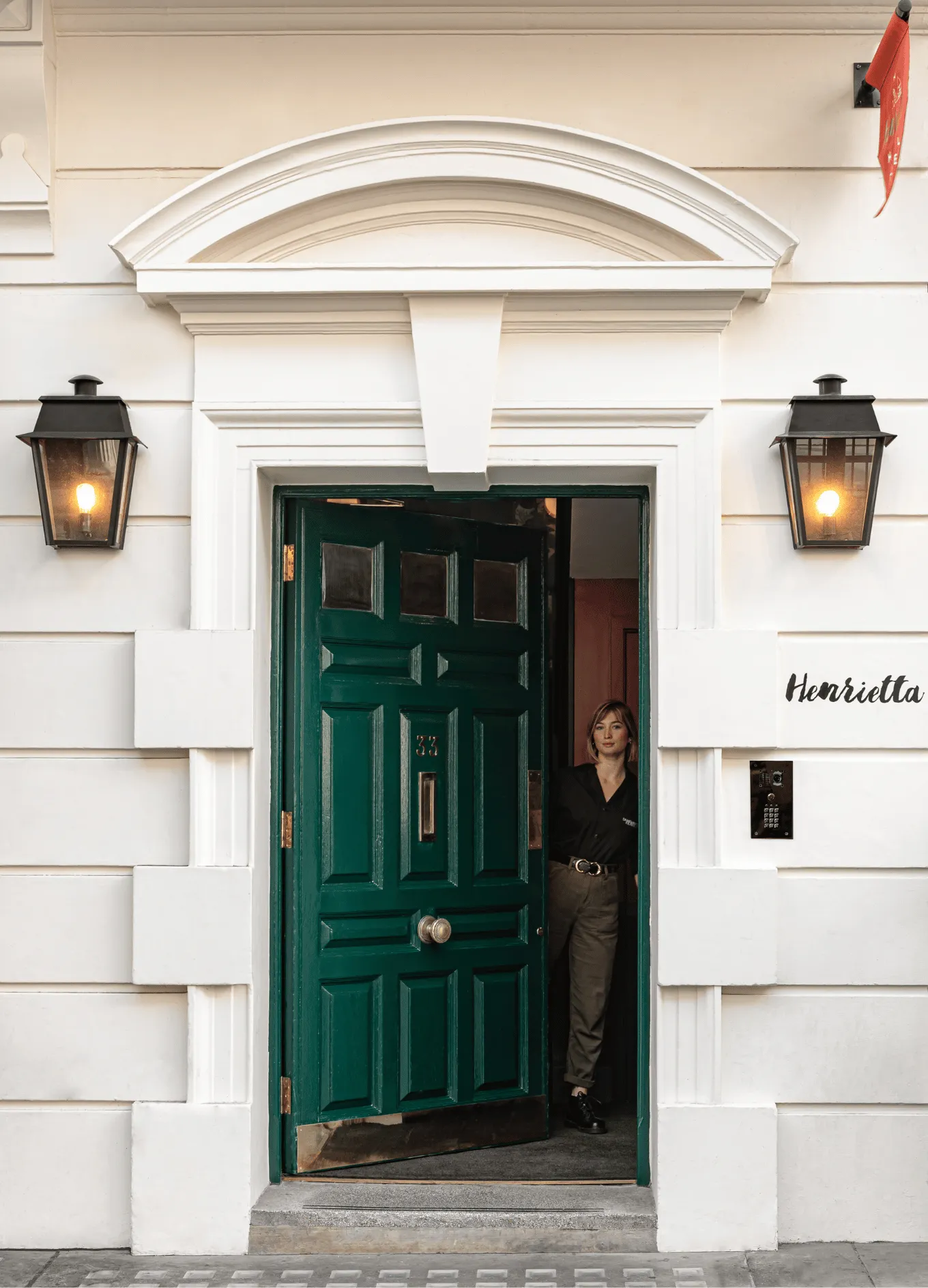 Woman standing in the doorway of a white building with a dark green door numbered 33 and sign reading Henrietta.
