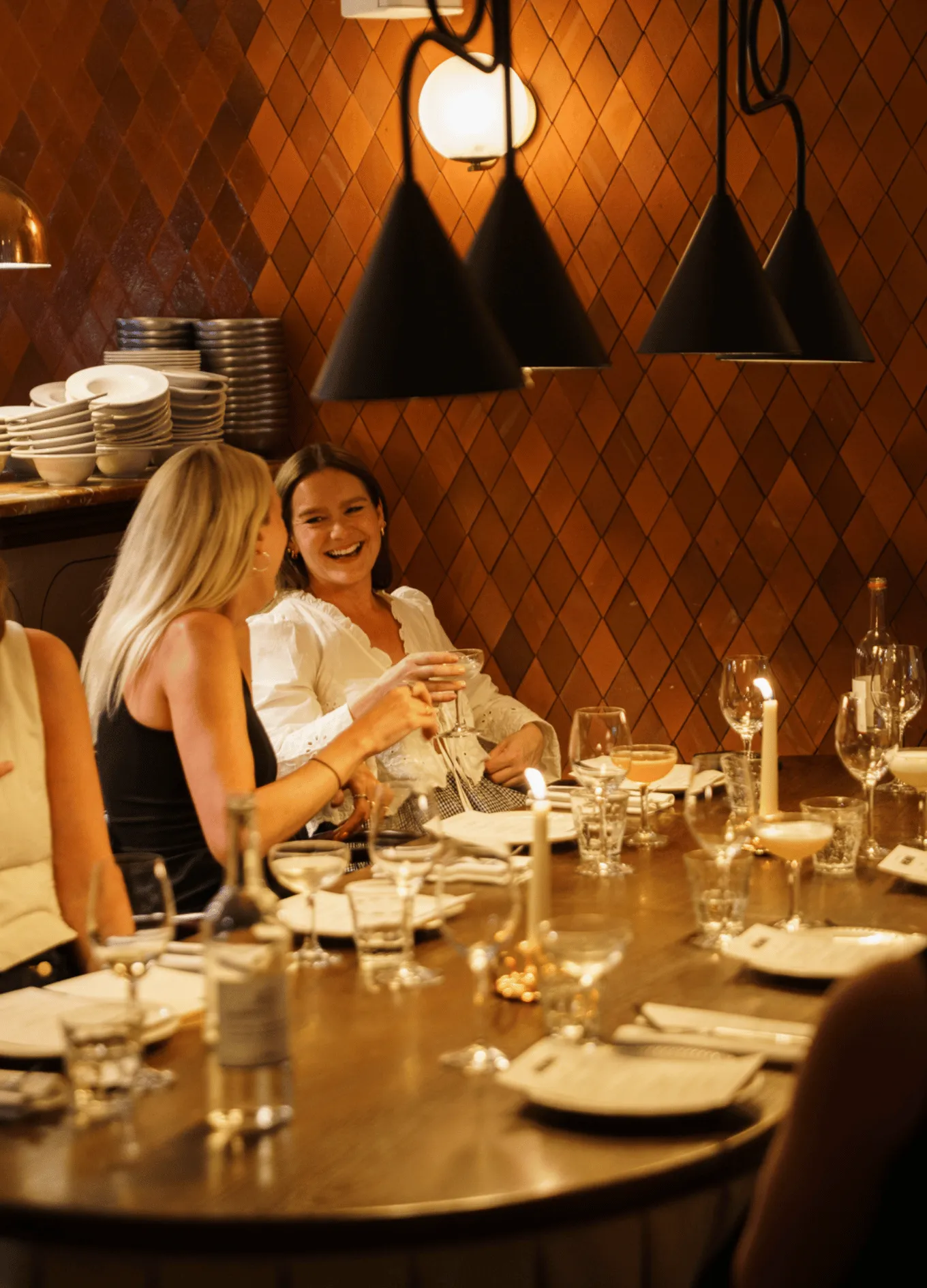 Two women laughing and enjoying drinks at a warmly lit restaurant table with candles and glassware.