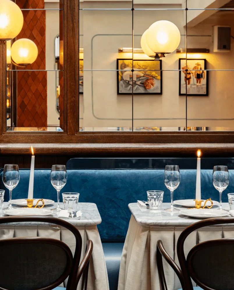 Elegant restaurant tables with white candles, empty wine glasses, and blue cushioned seating against a mirrored wall with framed artwork.