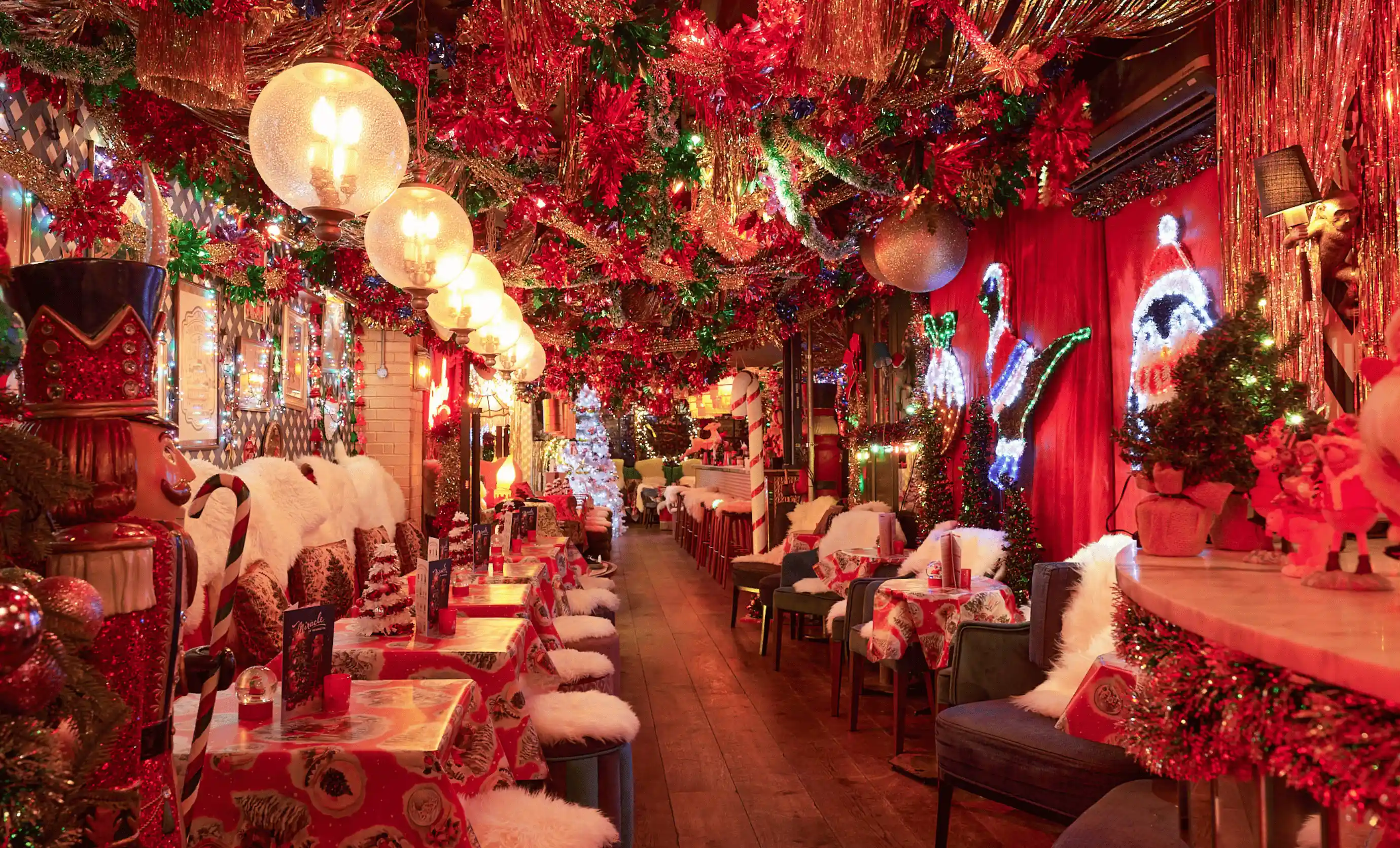 Interior of a festive restaurant decorated with red and green Christmas garlands, hanging lights, nutcracker figures, and tables covered with holiday-themed tablecloths and fluffy chair covers.