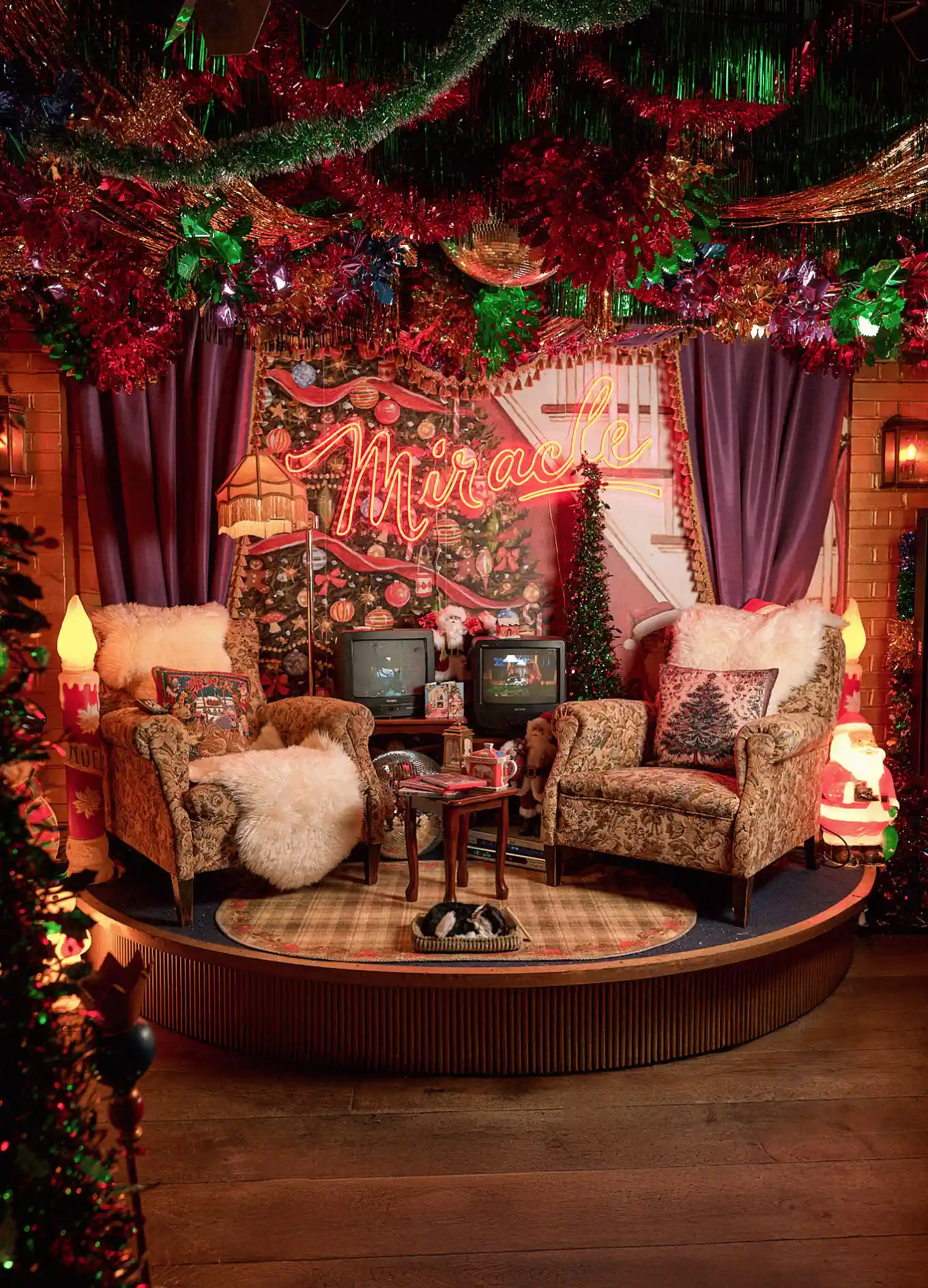 Cozy Christmas-themed sitting area with two upholstered armchairs, a small table, festive pillows, vintage TVs, and a neon sign reading 'Miracle'.
