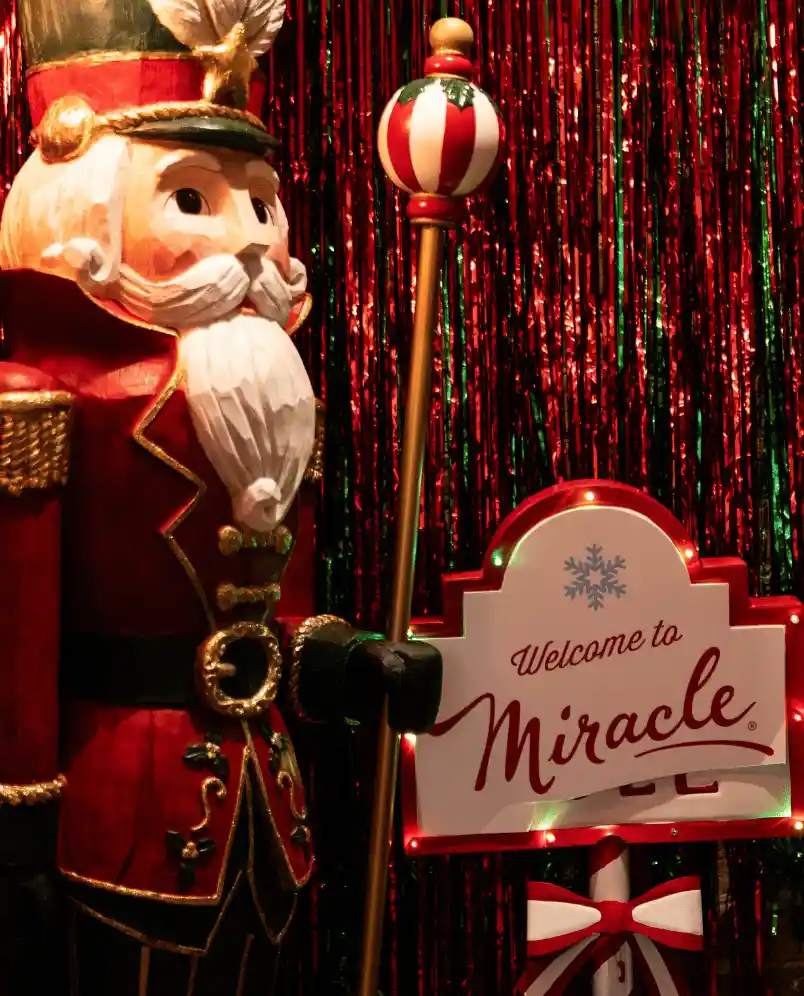 Wooden nutcracker soldier in red uniform holding a decorated staff next to a sign reading 'Welcome to Miracle' with festive red and green backdrop.