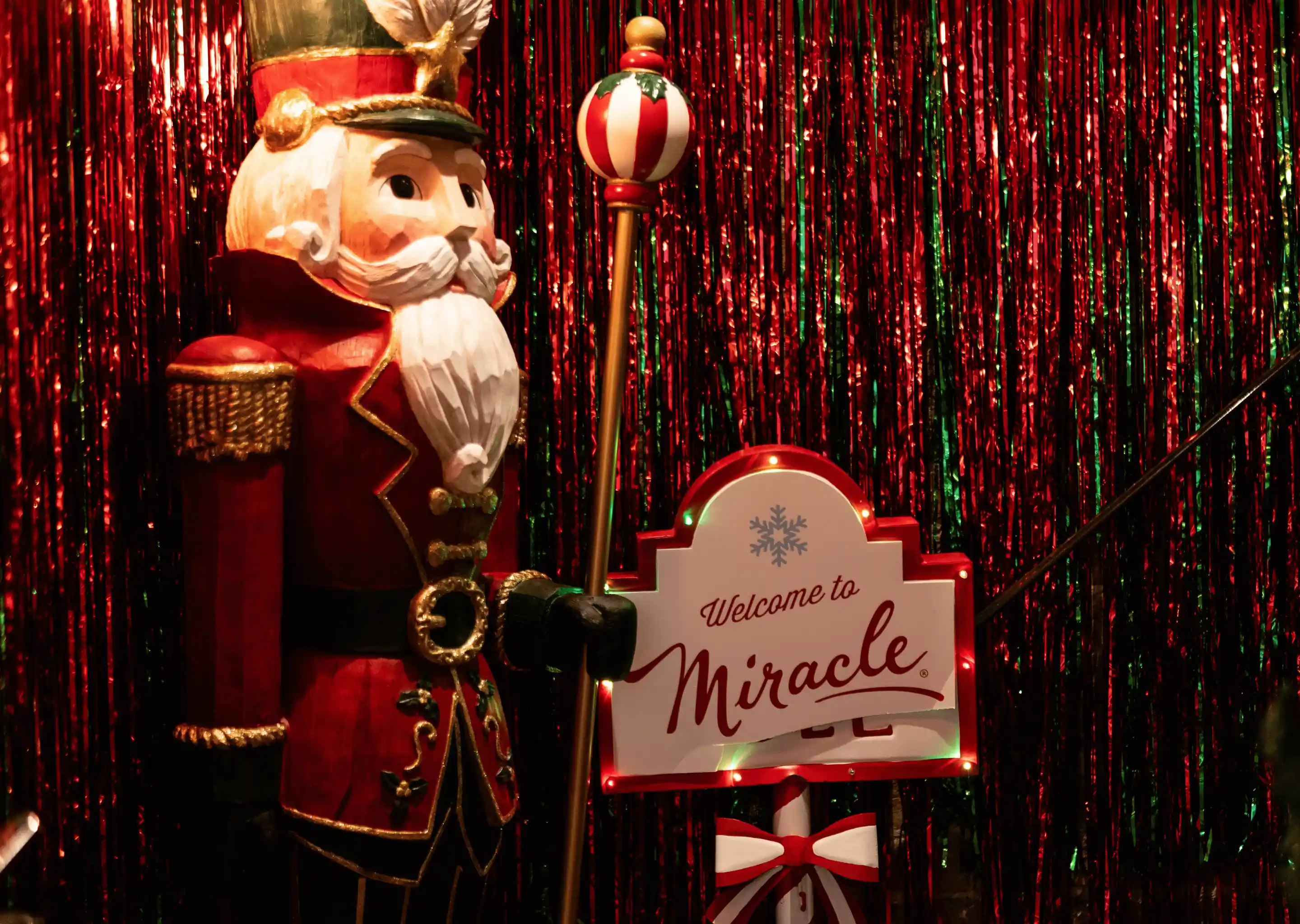 Decorative wooden nutcracker soldier in red uniform holding a staff next to a lit sign saying 'Welcome to Miracle' with red and green tinsel backdrop.