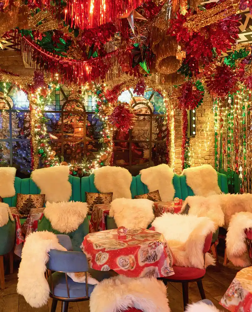 Festively decorated dining area with Christmas-themed tablecloths, chairs draped with white furry covers, and colorful lights and ornaments hanging from the ceiling.
