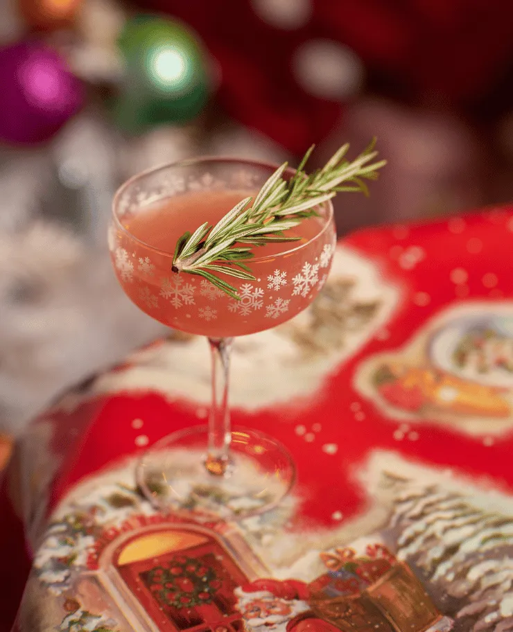 Festive pink cocktail in a snowflake-etched glass garnished with a sprig of rosemary on a Christmas-themed tablecloth.