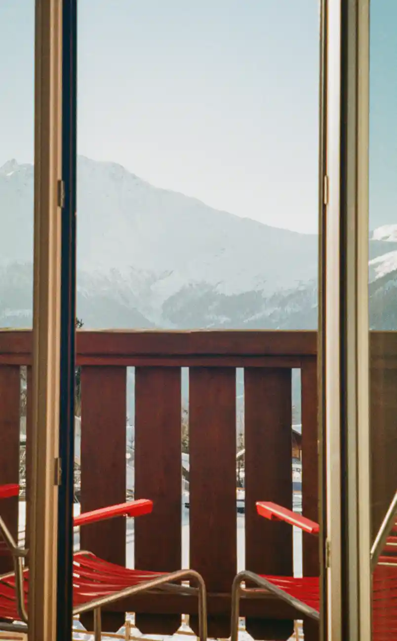 View through glass door of two red chairs on a balcony with snowy mountains in the background.