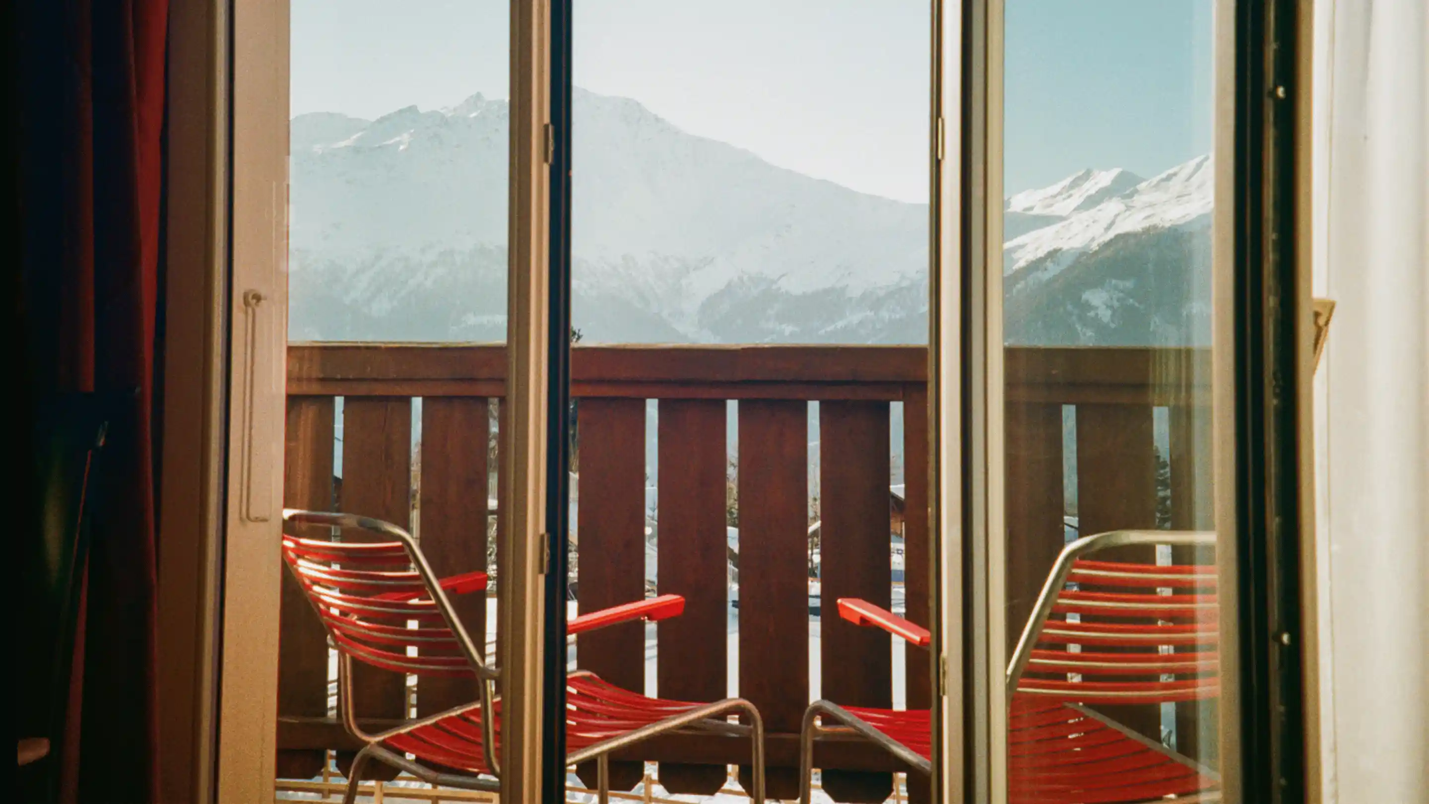 View through glass door of balcony with two red chairs and snowy mountain landscape in the background.