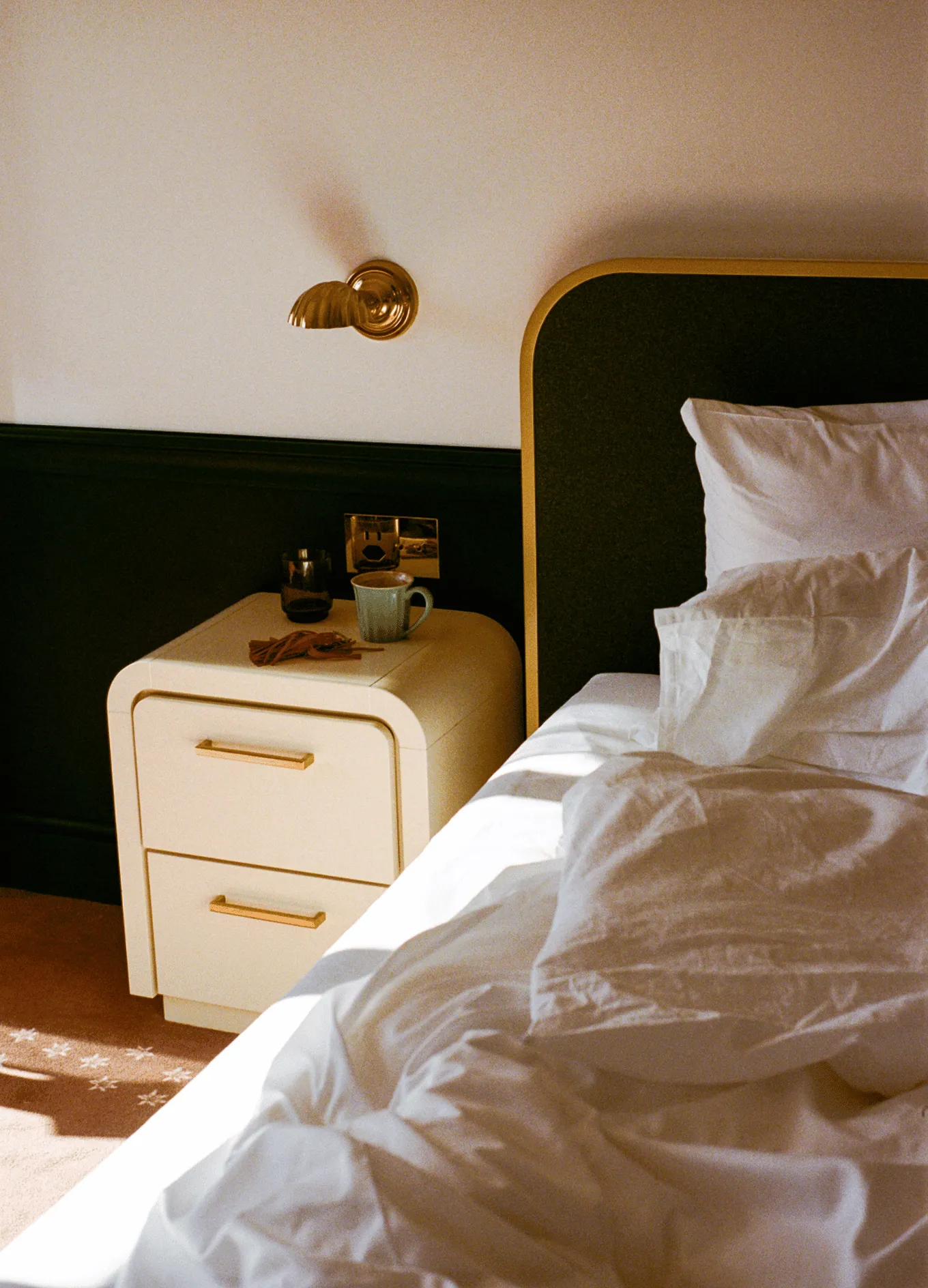 Acogedor rincón de dormitorio con cama sin hacer, sábanas blancas, una mesita de noche con una taza de café y llaves y una lámpara de pared dorada.