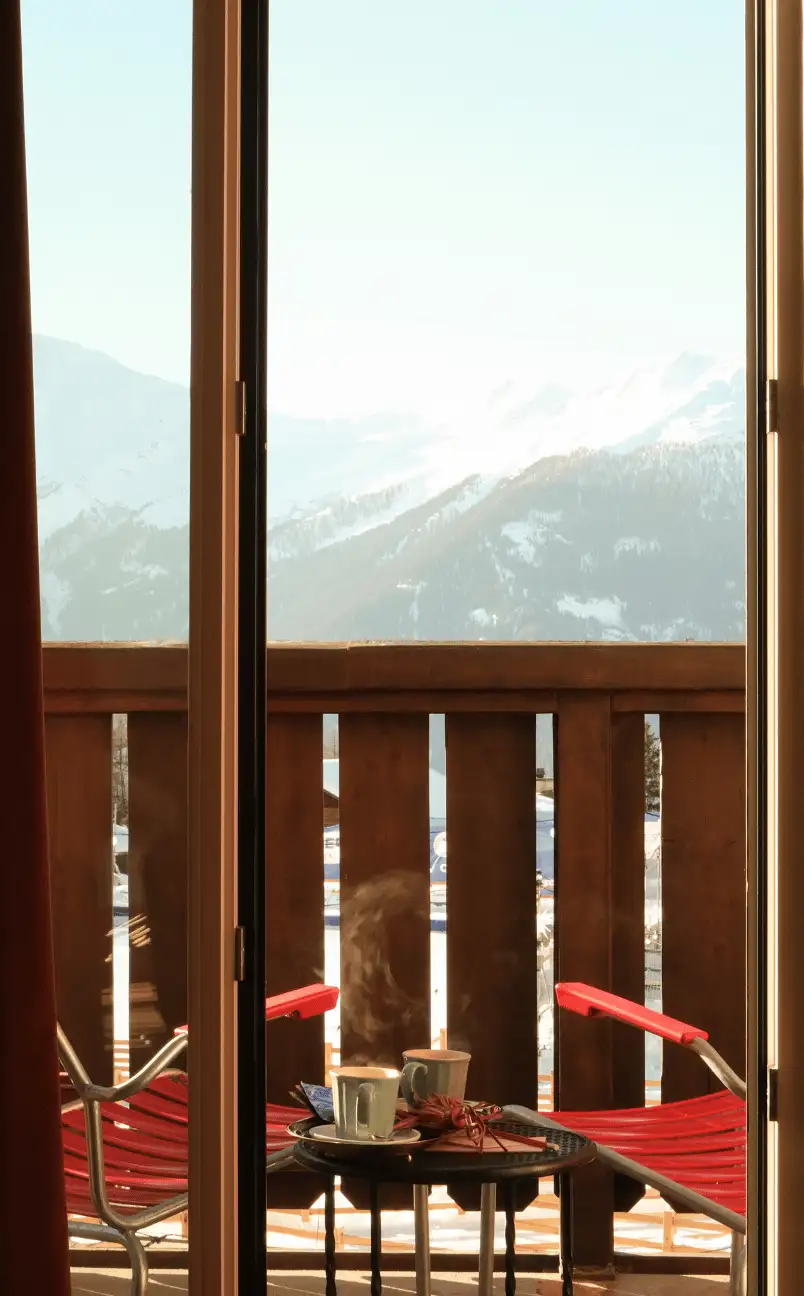 Vue à travers la porte d'un balcon montrant deux tasses fumantes posées sur une petite table et deux chaises rouges, avec un paysage de montagnes enneigées en arrière-plan.