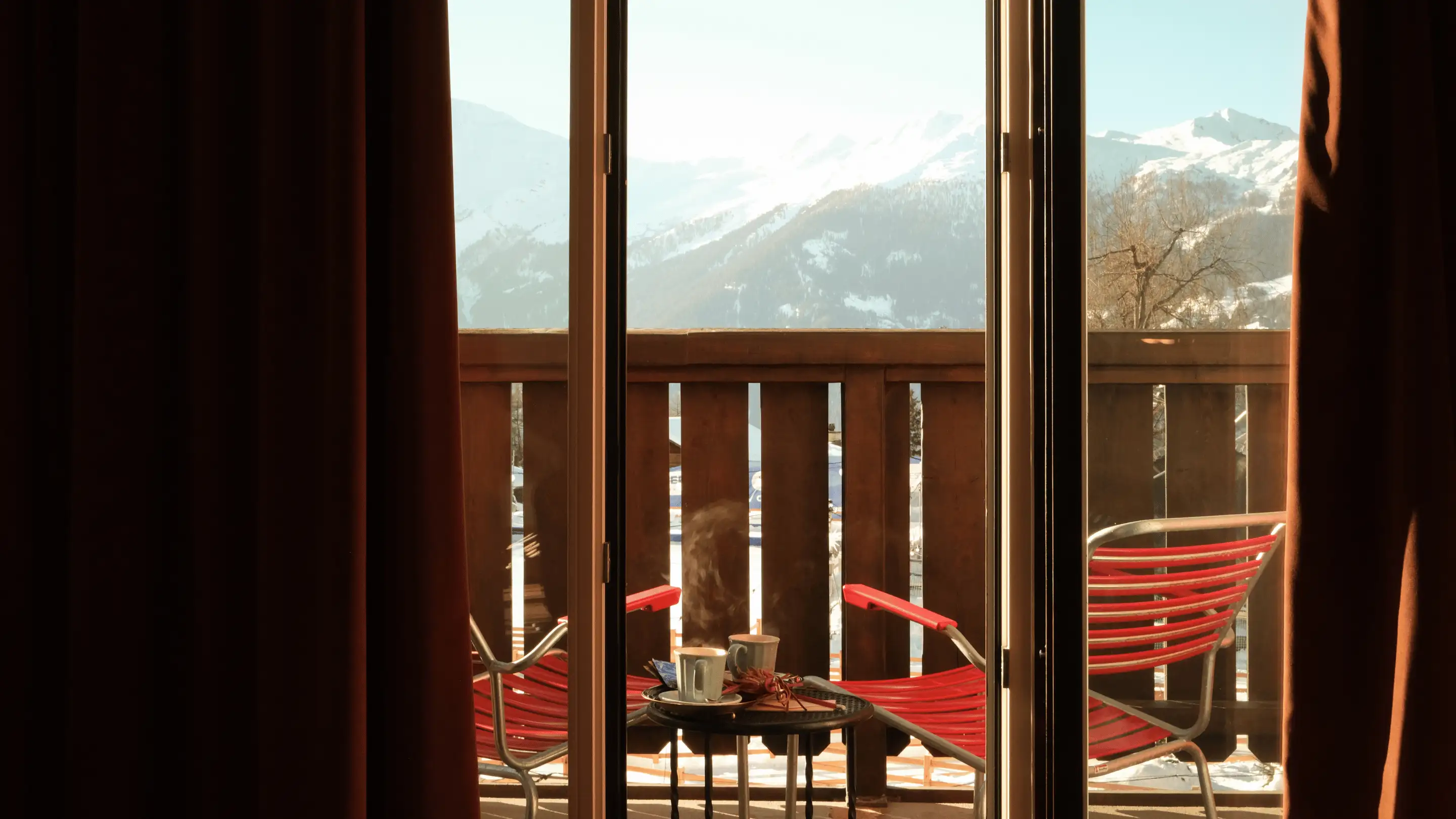 Vue à travers les portes vitrées d'un balcon avec deux chaises rouges et une petite table contenant des tasses fumantes, surplombant les montagnes enneigées.