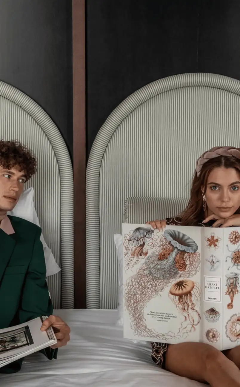 Un hombre y una mujer sentados en una cama con libros abiertos, y la mujer muestra una ilustración de medusas y vida marina.