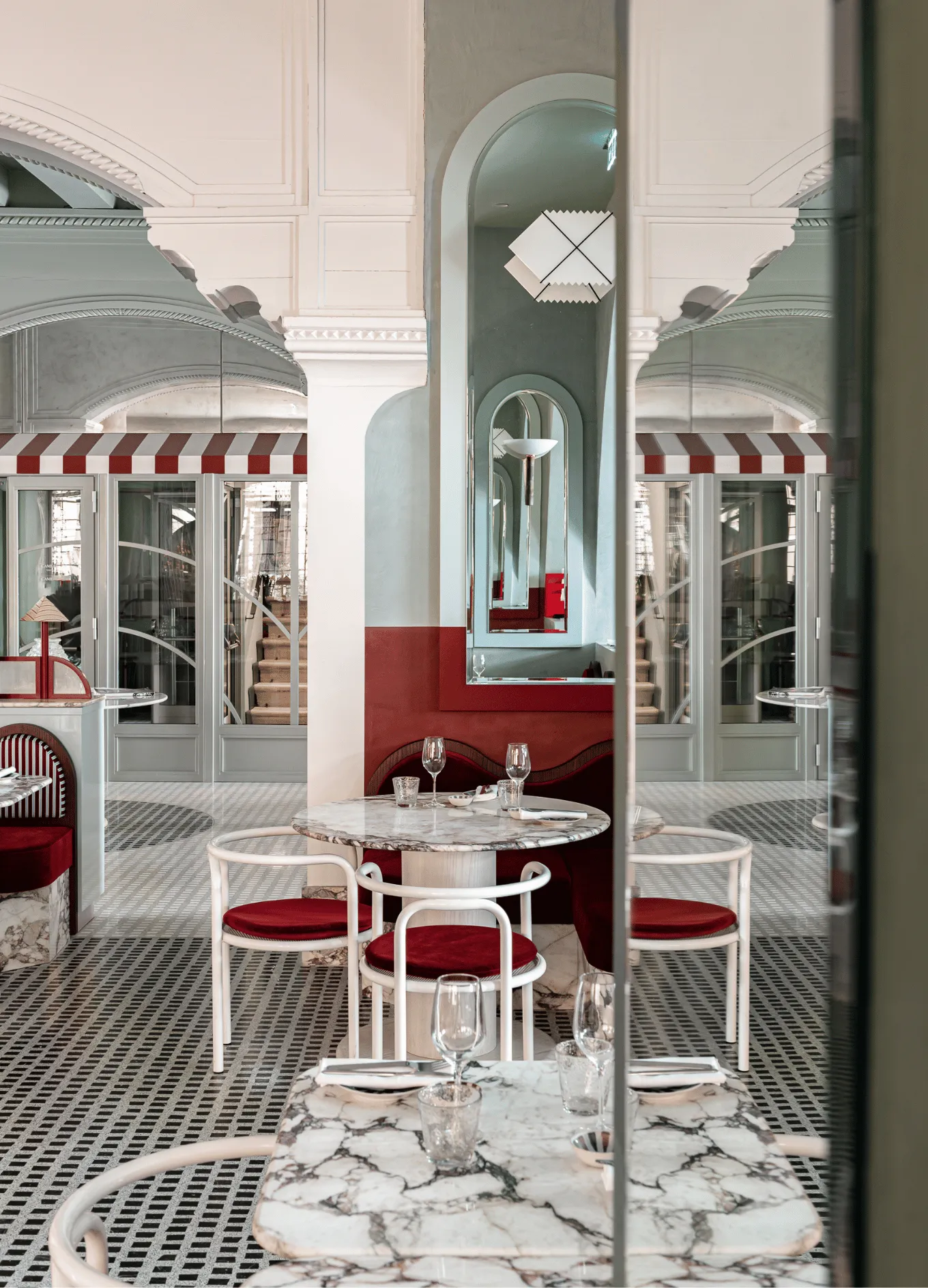 Elegant restaurant interior with marble tables, white chairs with red cushions, patterned floor, and arched mirrors reflecting the space.