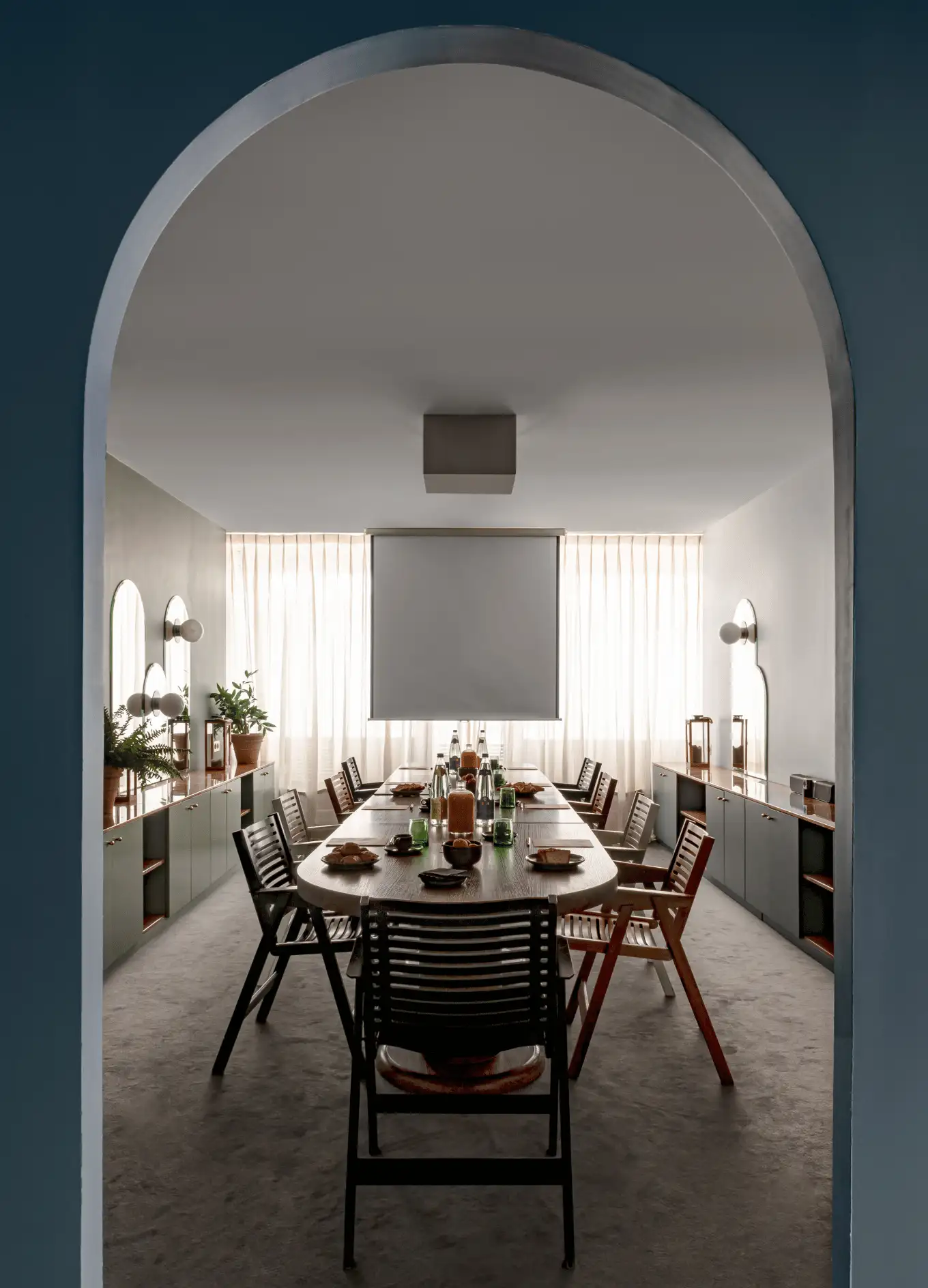 Sala de conferencias moderna y minimalista con una larga mesa de madera, sillas, plantas y una pantalla de proyección.