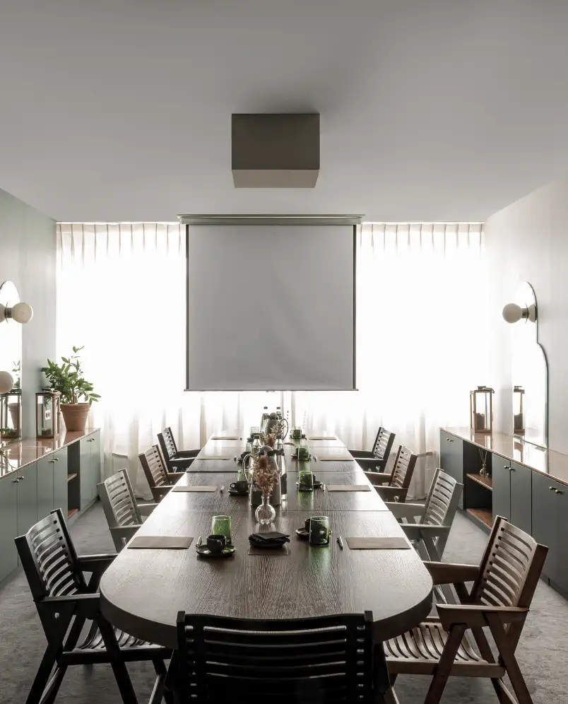 Sala de reuniones luminosa con una larga mesa de madera, varias sillas y una pantalla de proyección enrollable junto a ventanas con cortinas transparentes.
