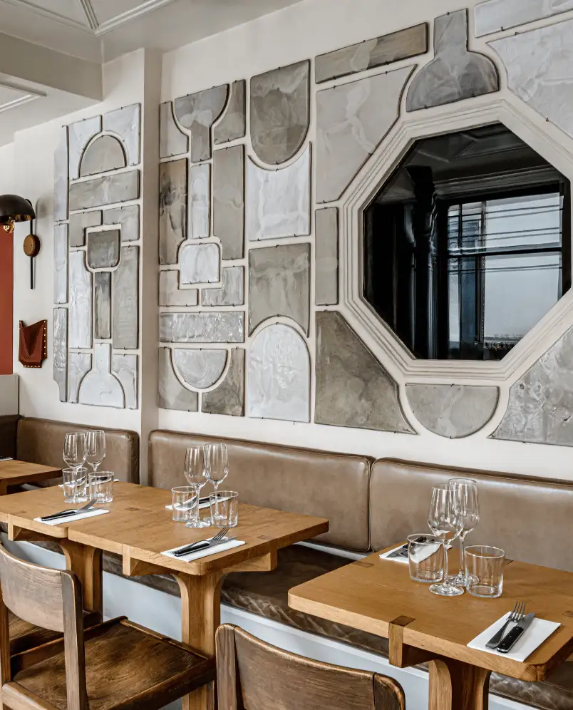 Área de estar del restaurante con mesas de madera, banco de cuero marrón, sillas de madera y decoración de pared abstracta con un espejo octogonal.