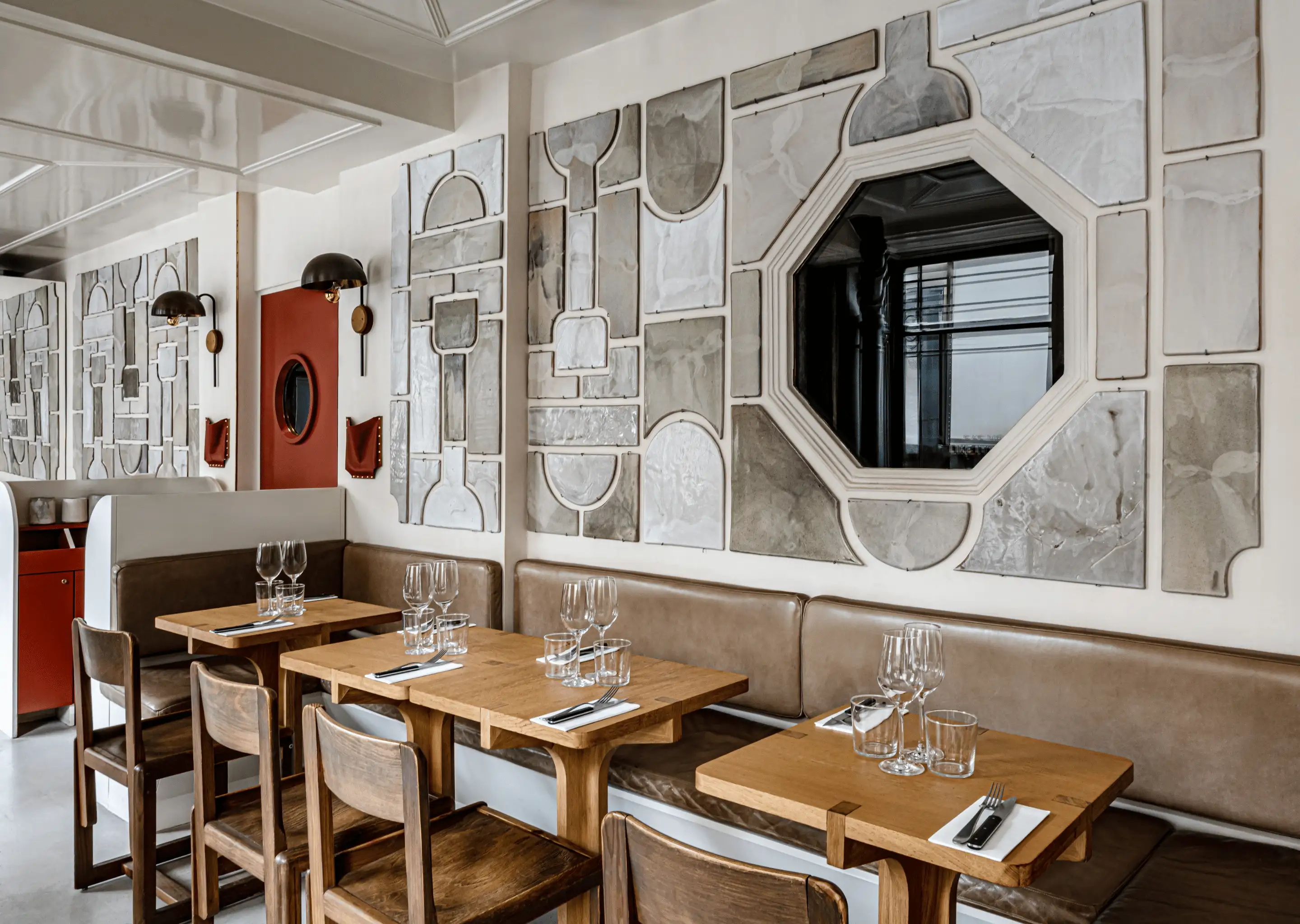 Acogedor interior de restaurante con mesas de madera, bancos de cuero marrón y arte mural abstracto con formas geométricas y un espejo octogonal.