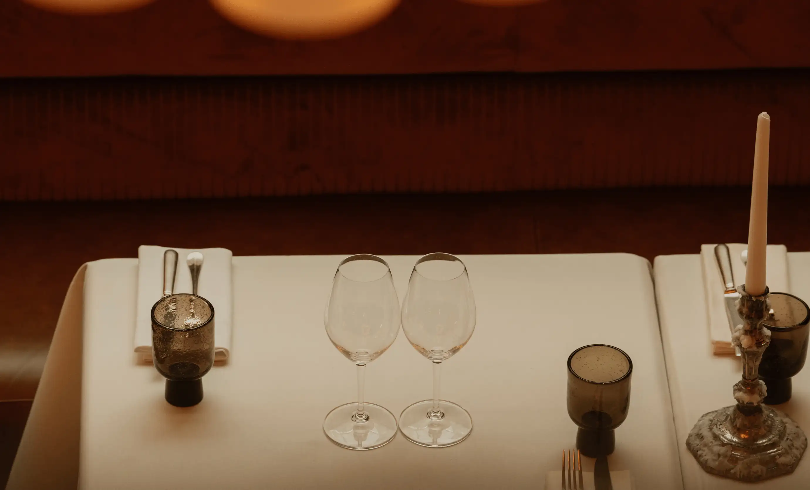 Table set for two with two wine glasses, dark tinted water glasses, silverware on white napkins, and a single tall candle in an ornate holder.